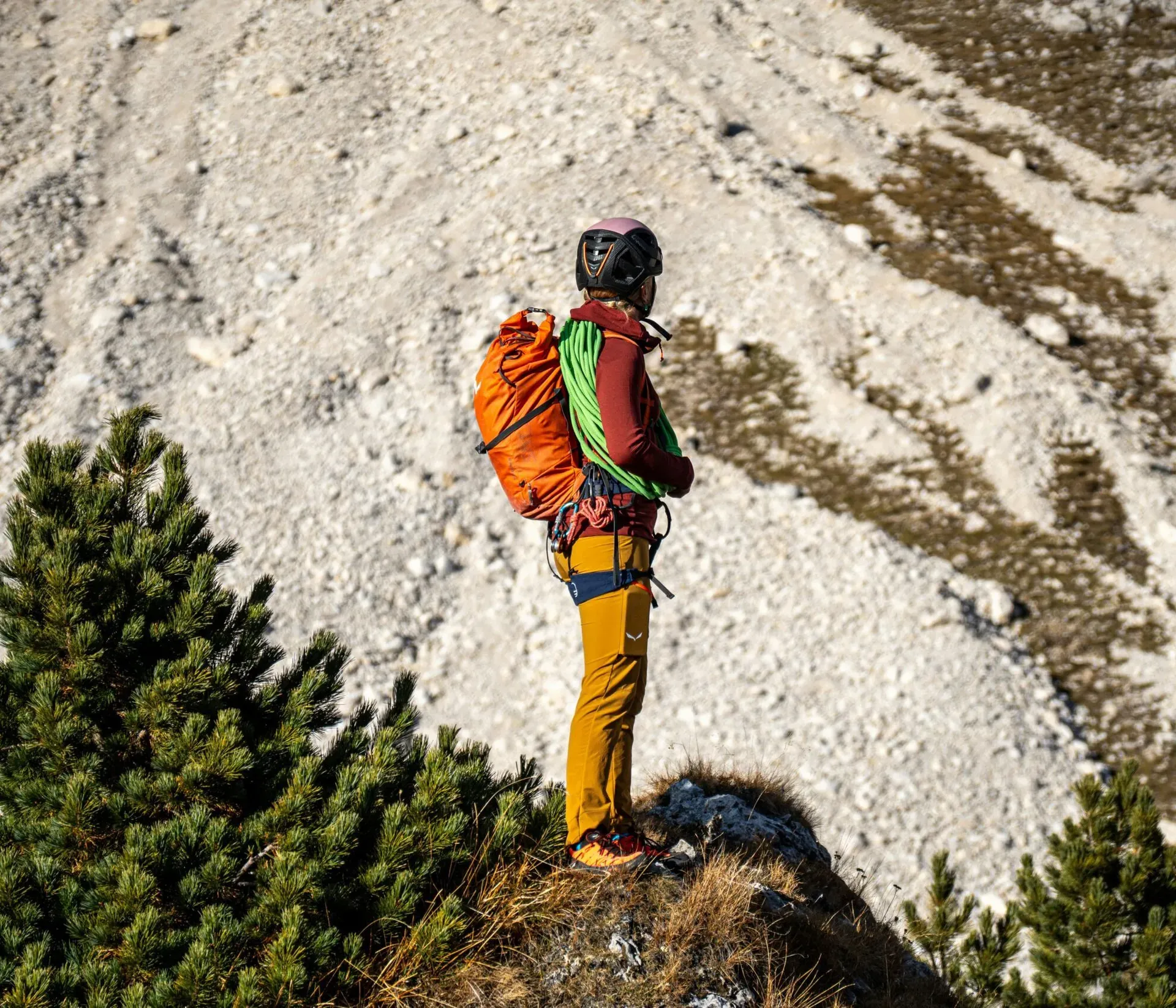 Hiker wearing a helmet, bright clothing, and an orange backpack standing on a grassy ledge, overlooking a sunlit rocky mountain landscape.