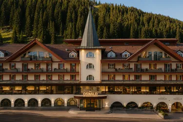 Exterior view of The Oswald Grand Hotel at sunset, featuring its distinctive central tower topped by a tall conical roof, elegant cream and wood façades, balconies and numerous illuminated windows. The main entrance opens onto the street, while a dense fir forest covers the mountain slope directly behind the hotel.
