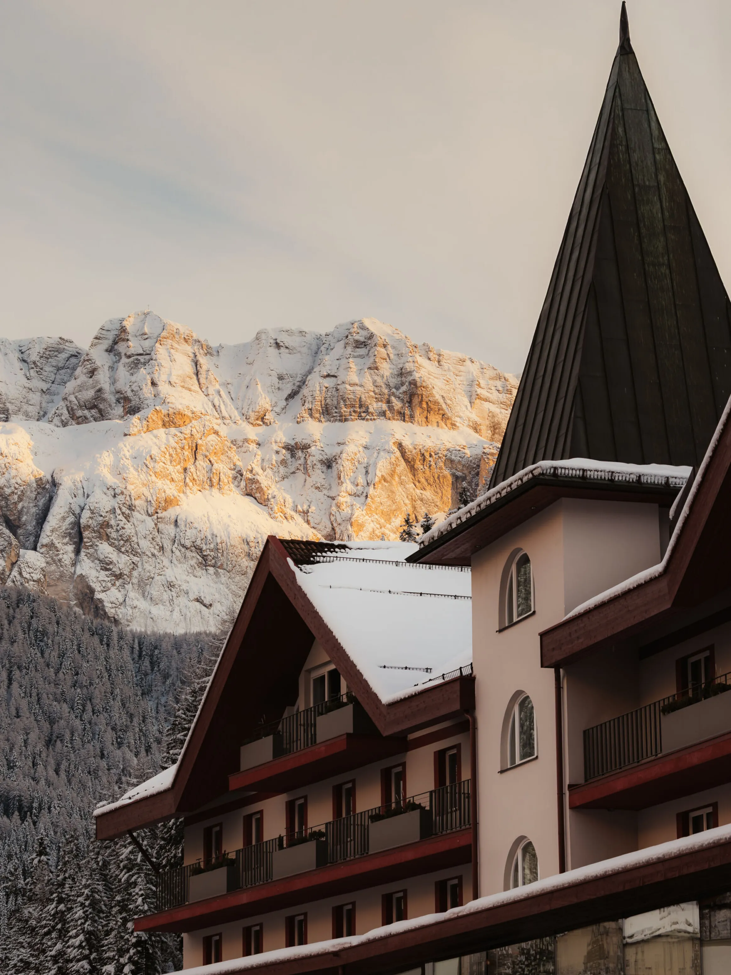Snow-covered alpine hotel with a steep roof sits in front of sunlit, snowy mountains. A tranquil, wintry atmosphere is captured at sunset.