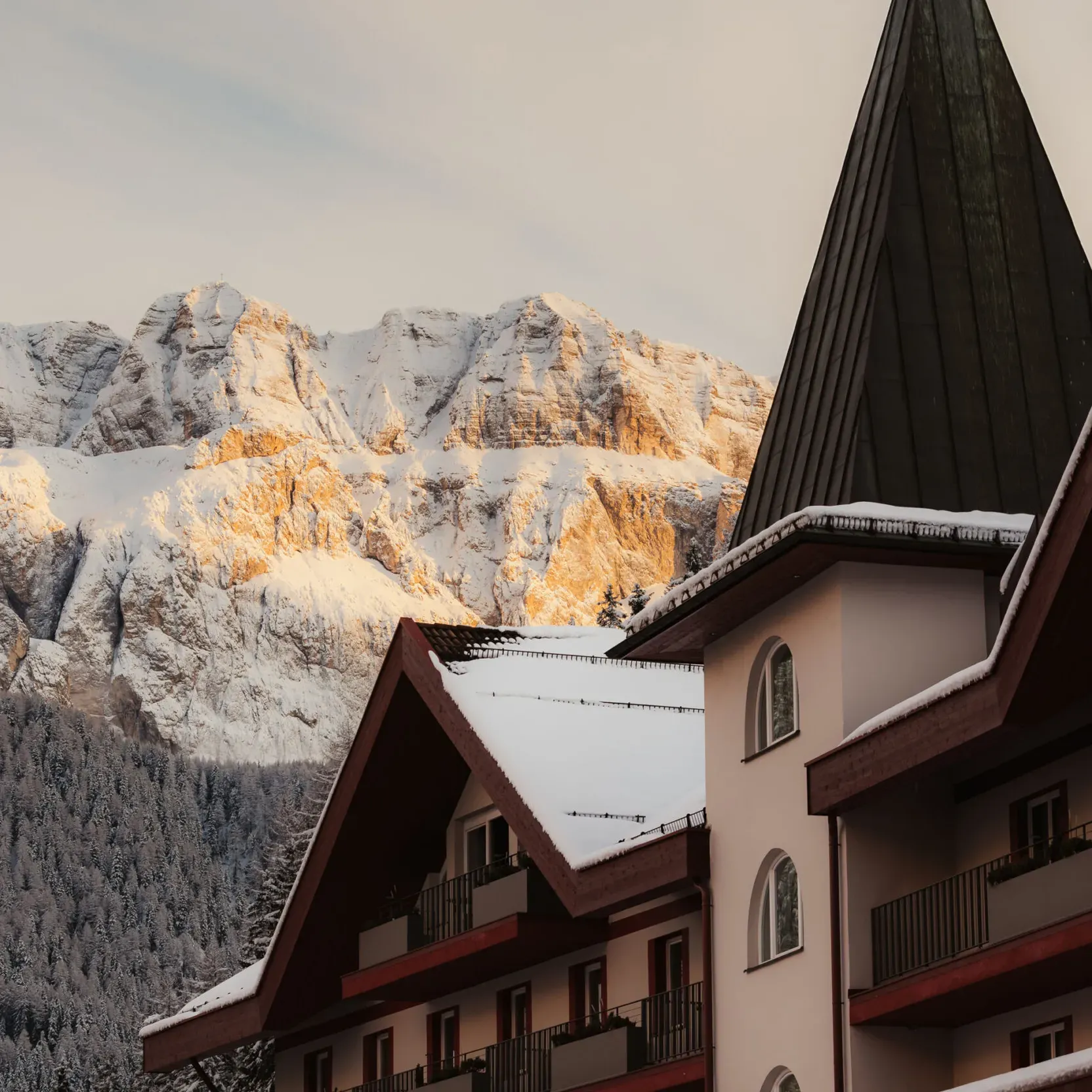 Snow-covered alpine hotel with a steep roof sits in front of sunlit, snowy mountains. A tranquil, wintry atmosphere is captured at sunset.