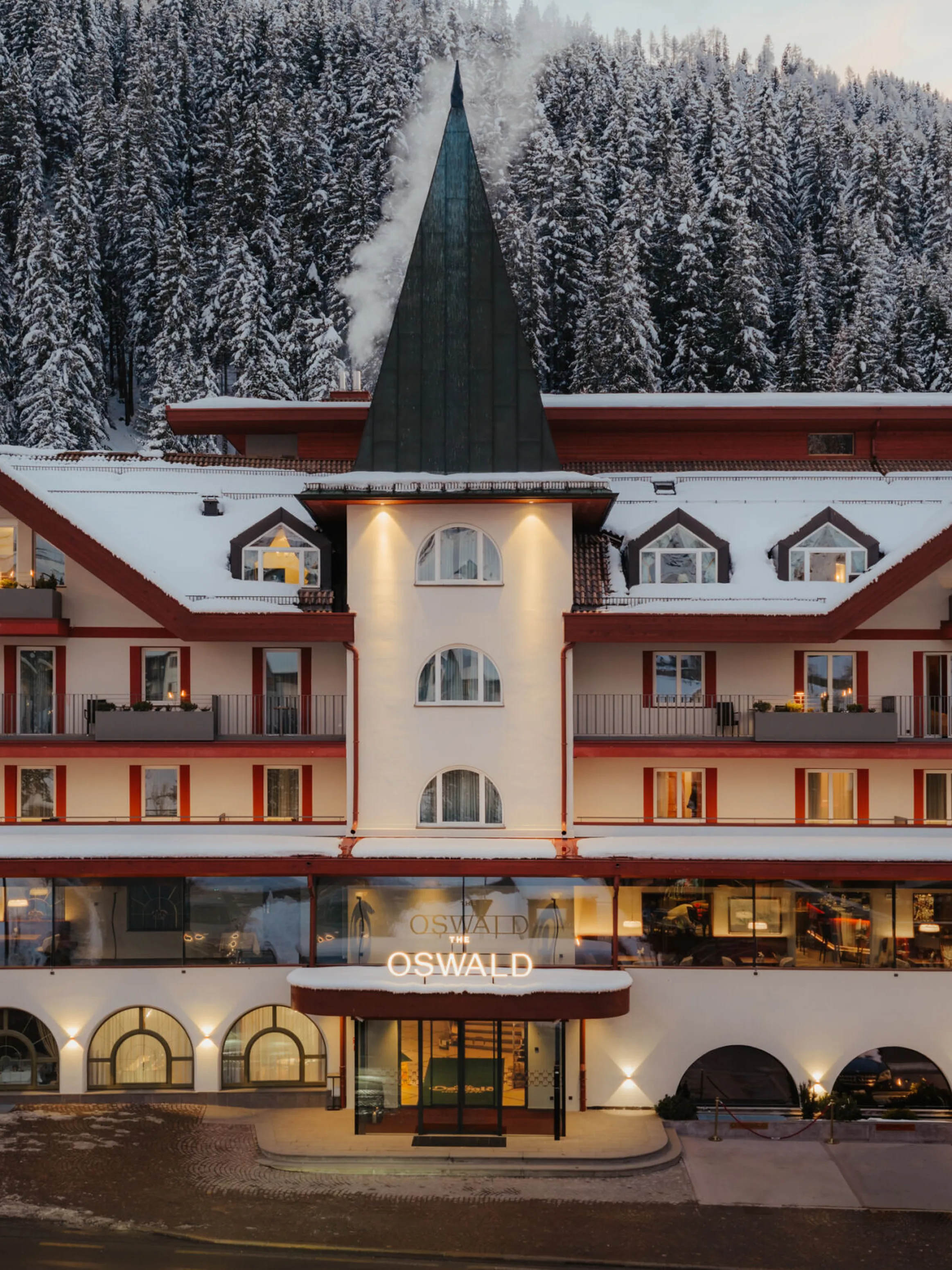 A cozy multi-story hotel with a central tower and snowy roof, set against a backdrop of snow-covered pine trees, exudes a warm, inviting ambiance.