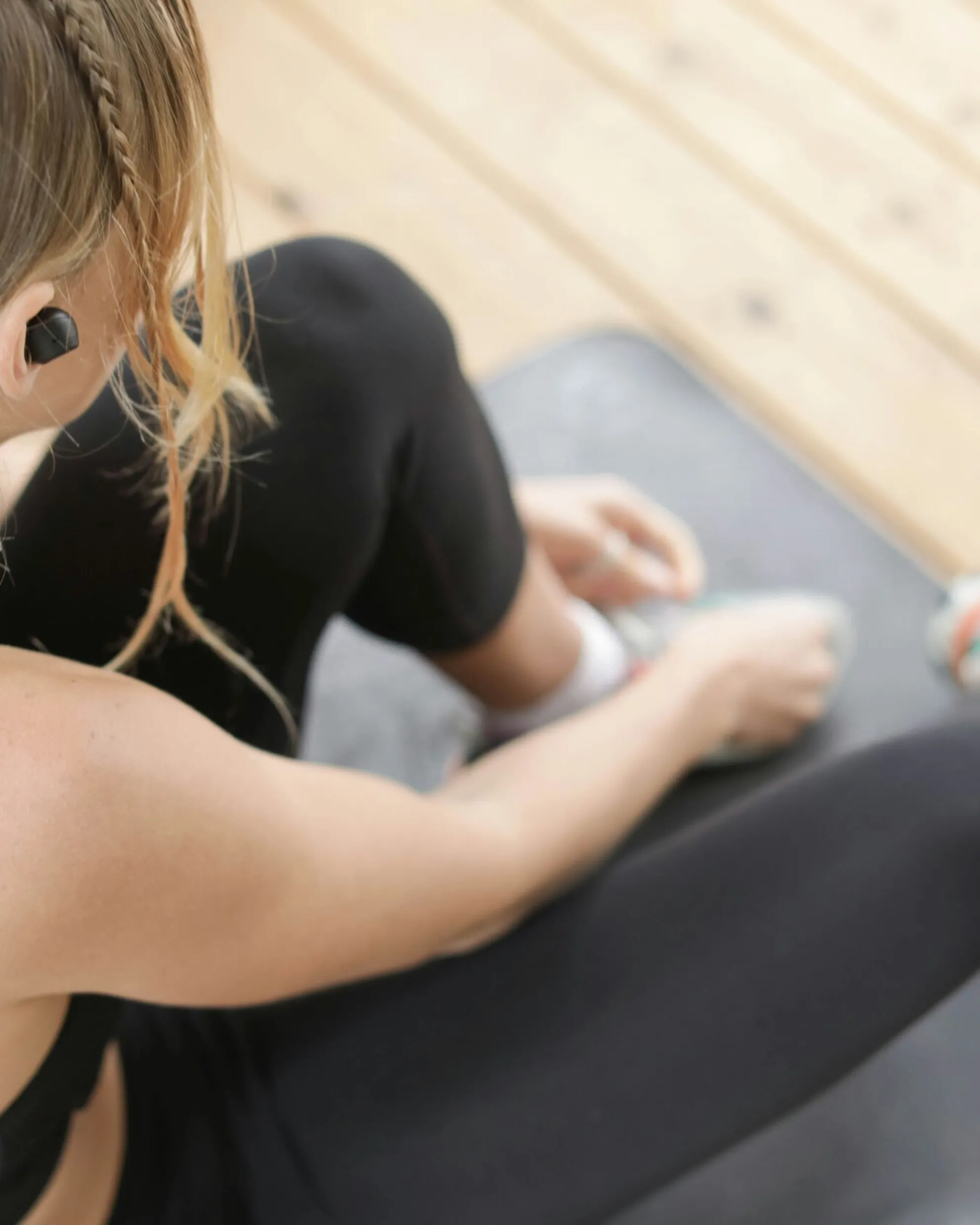 Blonde girl wearing earbuds and a black sports outfit, sitting on a fitness mat while tying her shoes before starting a bodyweight workout.