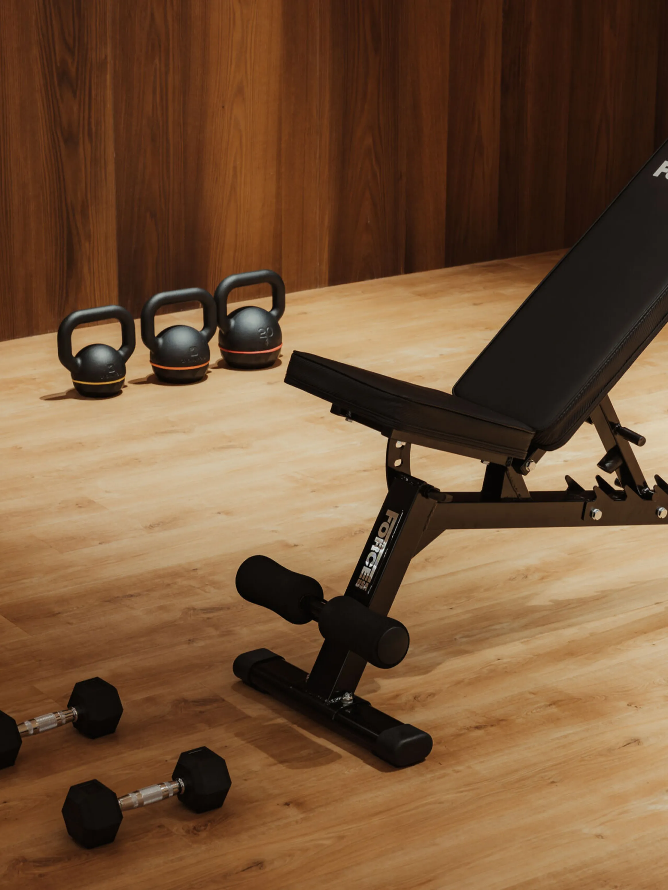 Adjustable black workout bench in a wooden home gym with kettlebells lined up on the floor and two dumbbells nearby. Warm, inviting atmosphere.