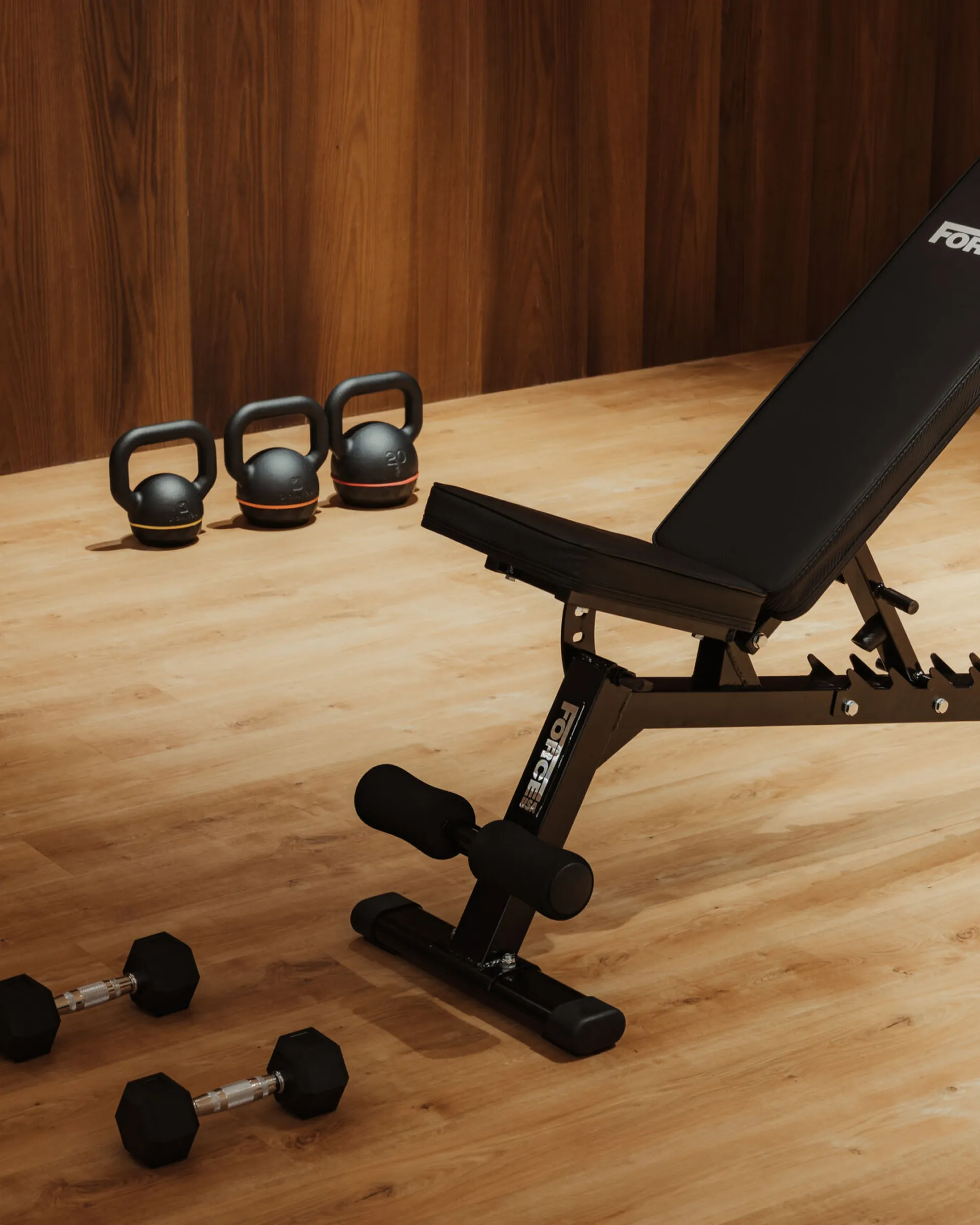 Adjustable black workout bench in a wooden home gym with kettlebells lined up on the floor and two dumbbells nearby. Warm, inviting atmosphere.