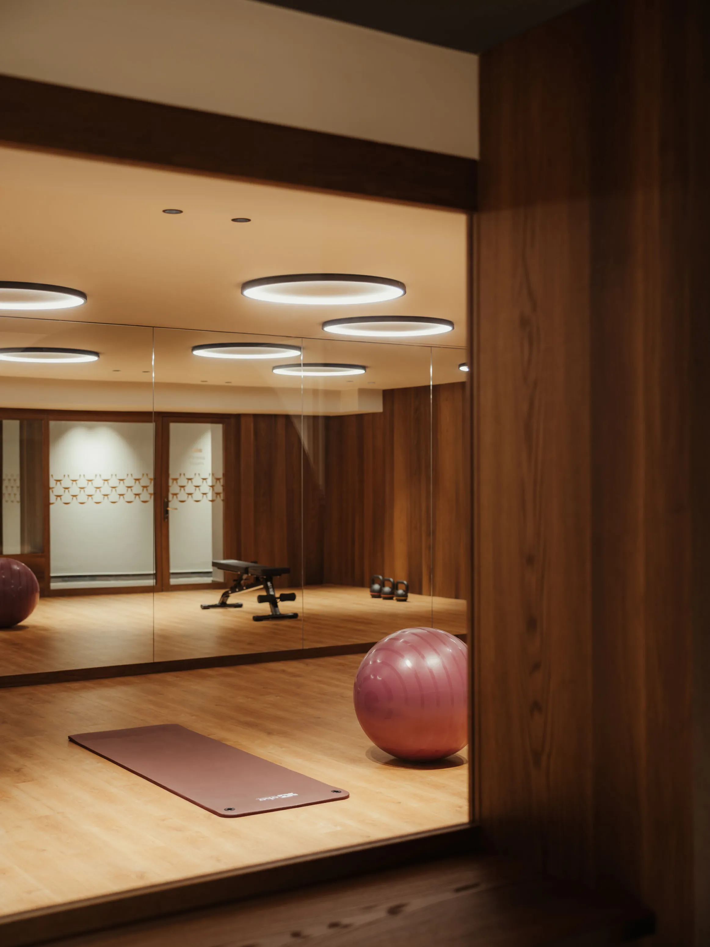 Modern gym with wooden walls and flooring, featuring a yoga mat, exercise ball, and mirrored walls reflecting sleek round ceiling lights.