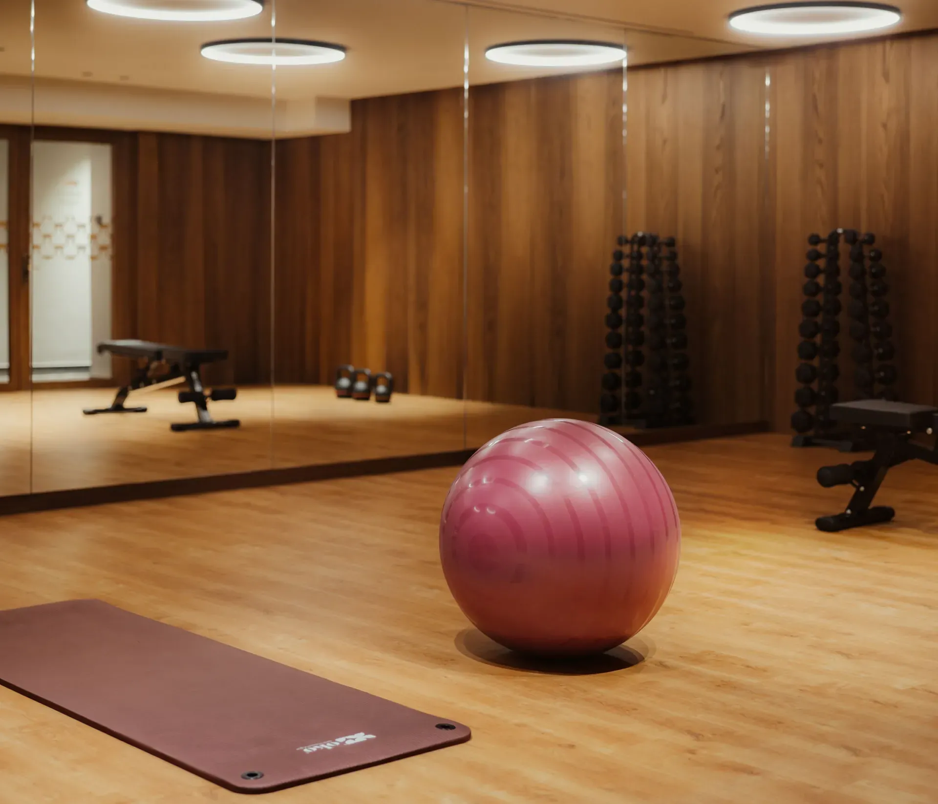 Ruhiger Fitnessraum mit Holzboden, einem großen lila Gymnastikball und einer bordeauxfarbenen Matte. Hanteln und Bänke spiegeln sich in den Spiegelwänden und werden warm beleuchtet.