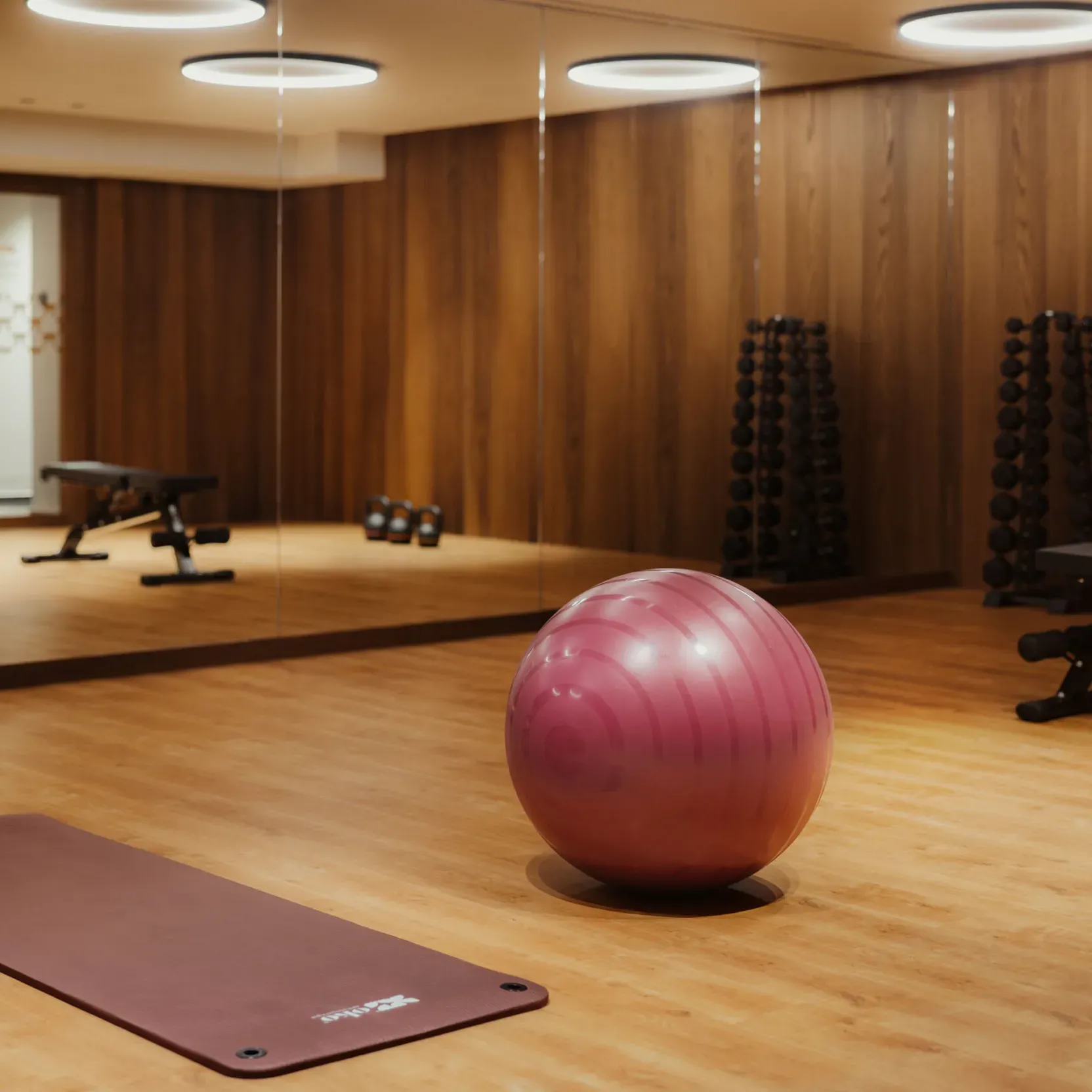 A serene gym with wooden floors, a large purple exercise ball, and a burgundy mat. Dumbbells and benches are reflected in mirrored walls, lit warmly.