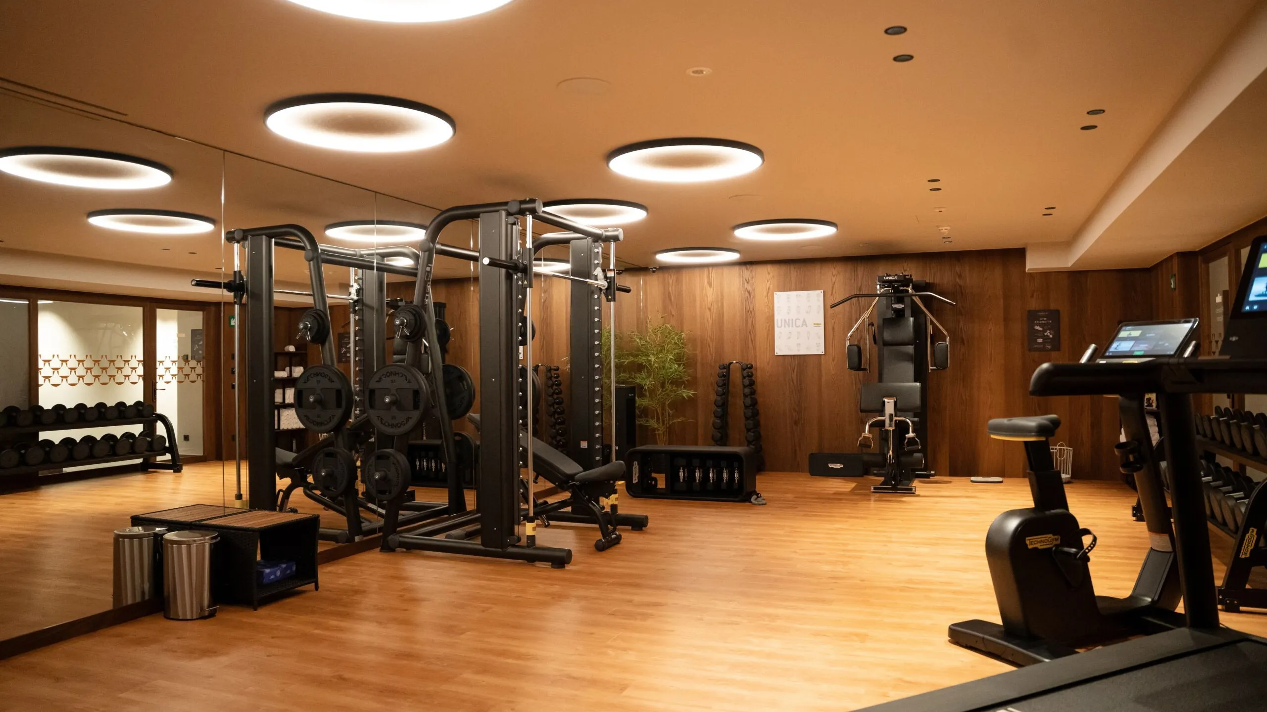 Spacious fitness area at The Oswald Grand Hotel, equipped with modern training equipment and featuring extensive natural wood finishes. The room includes various fitness and strength-training machines, light wooden flooring and warm indirect lighting. Large windows and mirrors create an open, bright training environment with an alpine-inspired atmosphere.