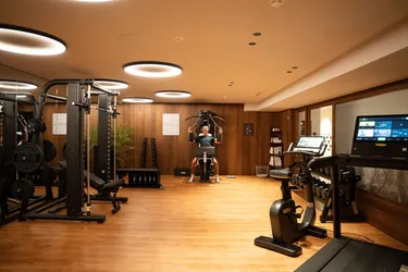 Panoramic view of the elegant fitness area at the Oswald Grand Hotel, featuring a sophisticated interior with natural wood flooring, wood-panelled walls and warm, atmospheric lighting. The gym is equipped with modern Technogym equipment for cardiovascular and strength training, including treadmills, cross trainers, exercise bikes, multifunction machines, benches, barbells and dumbbells. Large glass surfaces and mirrors enhance the sense of space, while the carefully arranged equipment creates an exclusive, refined and highly functional environment for guests.