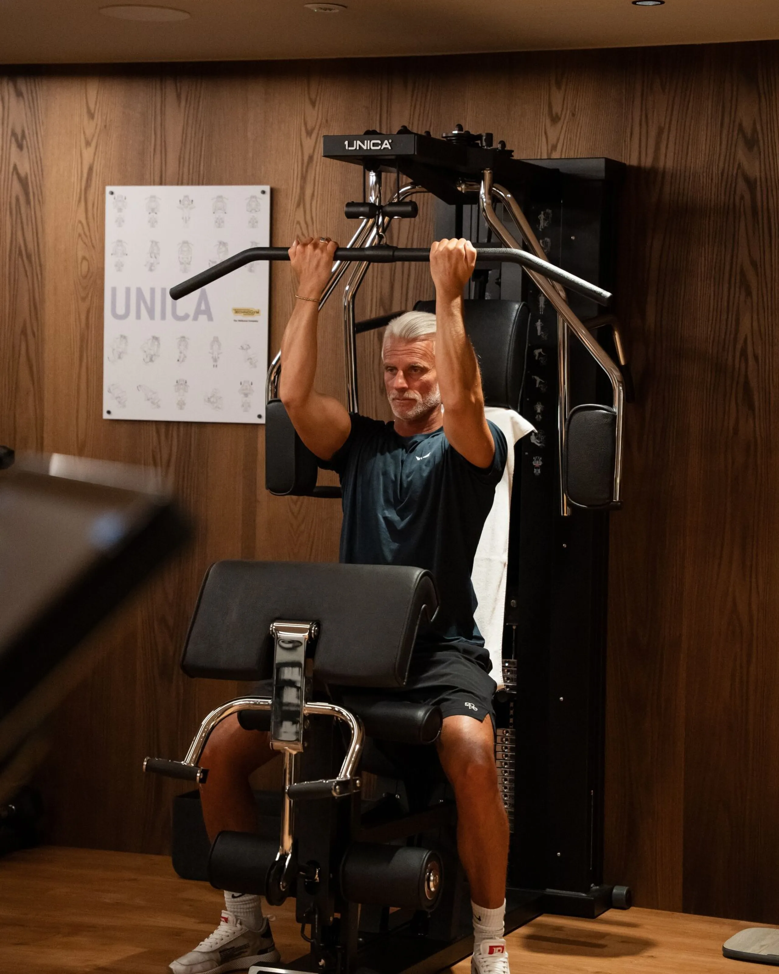 Oswald Grand Hotel guest performing a lat pulldown exercise on a Technogym training machine, seated at the station with both hands gripping the overhead bar during a controlled pulling movement. The multifunction machine occupies the centre of the image and is set within an elegant interior featuring natural wood-panelled walls, wooden flooring and warm ambient lighting. A decorative graphic and additional gym equipment can be seen in the background, highlighting the variety of training options available to hotel guests.