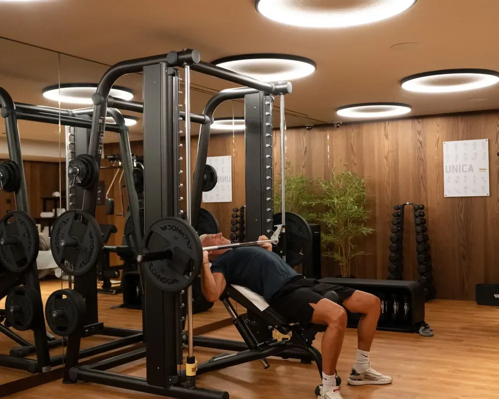 Oswald Grand Hotel guest during a strength-training session, performing a barbell exercise on an incline bench inside the Technogym fitness area. The multifunction training station and weight-training structure are positioned around the bench, with additional weights and professional equipment visible in the background. Wood-panelled walls and the warm glow of the circular ceiling lights create an elegant and intimate setting dedicated to fitness, wellbeing and active relaxation.