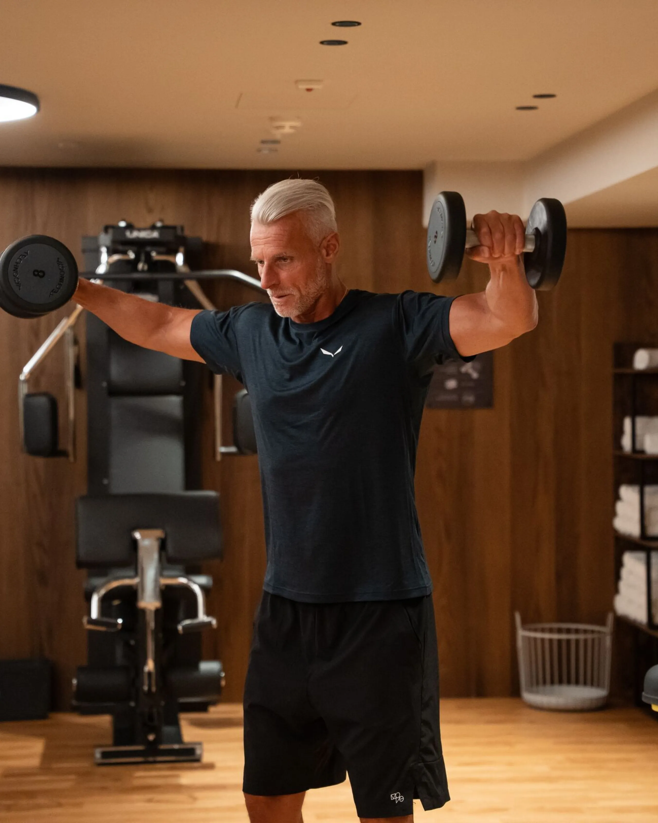 Oswald Grand Hotel guest performing a lateral raise with two dumbbells in the hotel fitness area. The guest is standing in the centre of the training space with both arms extended to the sides, while professional Technogym machines, a weight rack and the distinctive wood-panelled walls can be seen in the background. Warm ambient lighting and clean interior lines create a modern, elegant setting focused on movement, fitness and wellbeing.