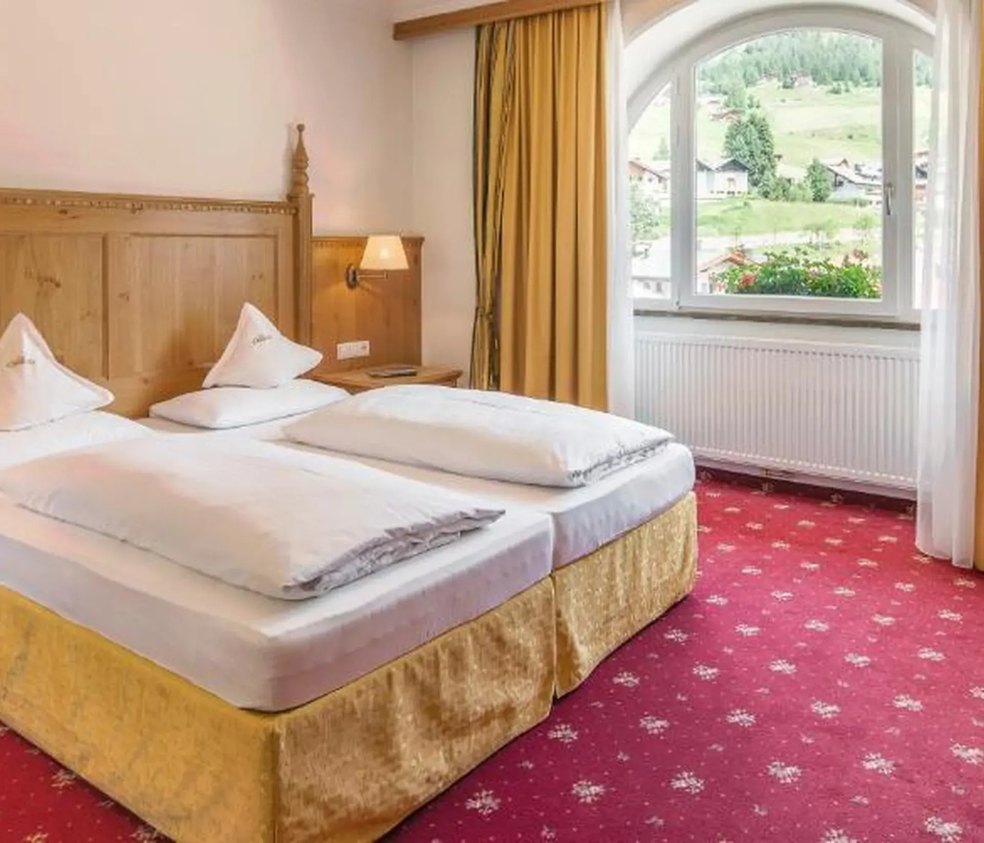 Historic room at Hotel Oswald with Swiss pine wood, red carpet, draped yellow fabric curtains, bedside lamps on, and a view of the forest.