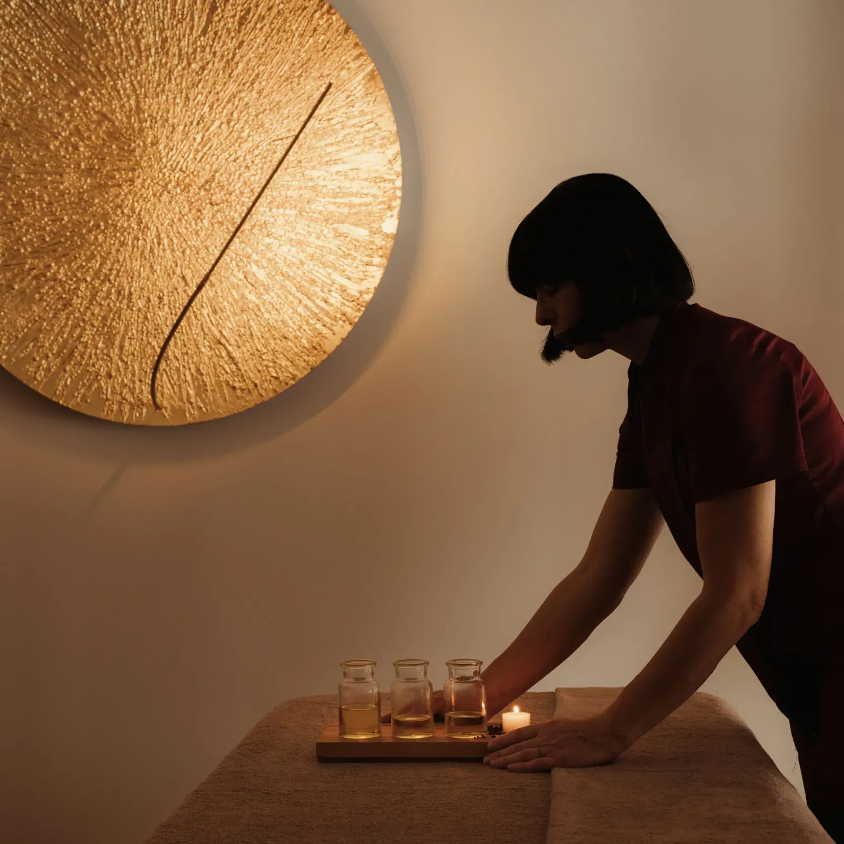 Una persona in una spa posiziona un vassoio con piccoli bicchieri di vetro su un lettino da massaggio. Una grande lampada circolare dorata e dalla superficie strutturata adorna la parete, diffondendo una luce calda.