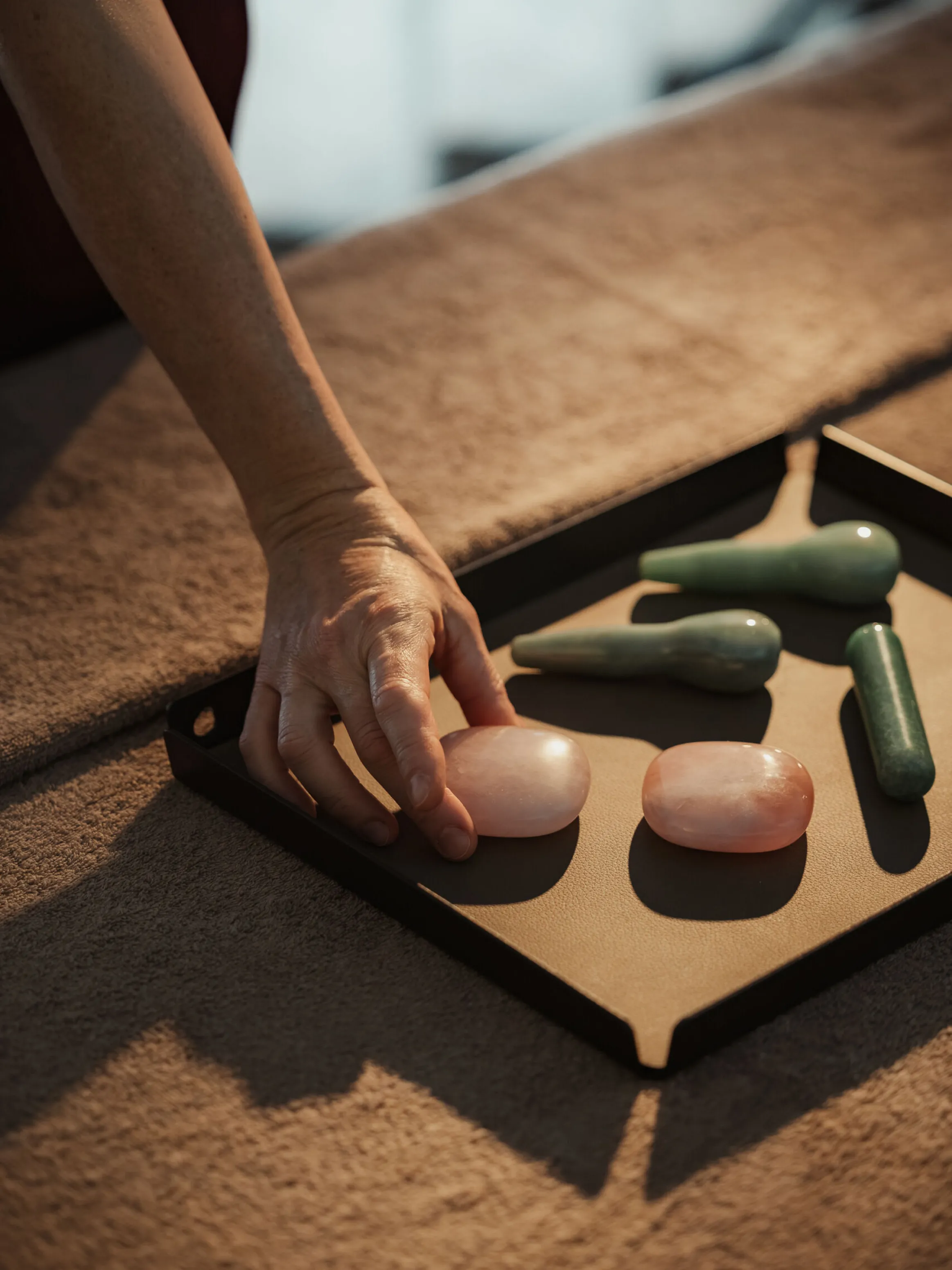 A hand reaches for a smooth, pink stone on a tray, surrounded by other green massage stones, on a soft, brown surface. The setting feels calm and soothing.
