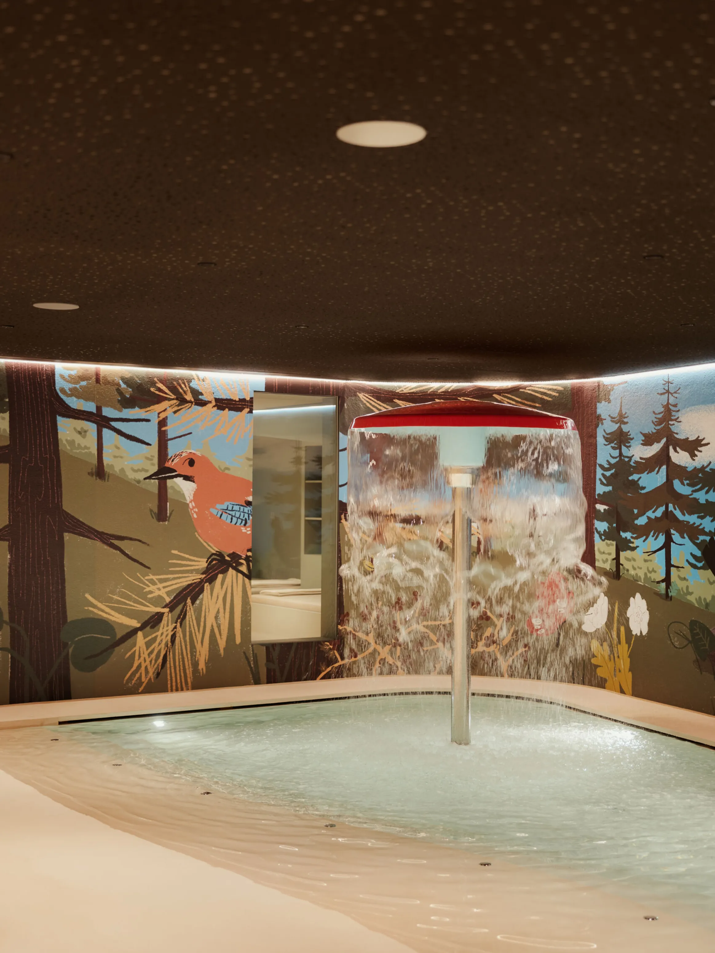 Indoor pool area with a red mushroom-shaped water feature splashing water. The walls display murals of trees and a bird, creating a playful, nature-inspired setting.