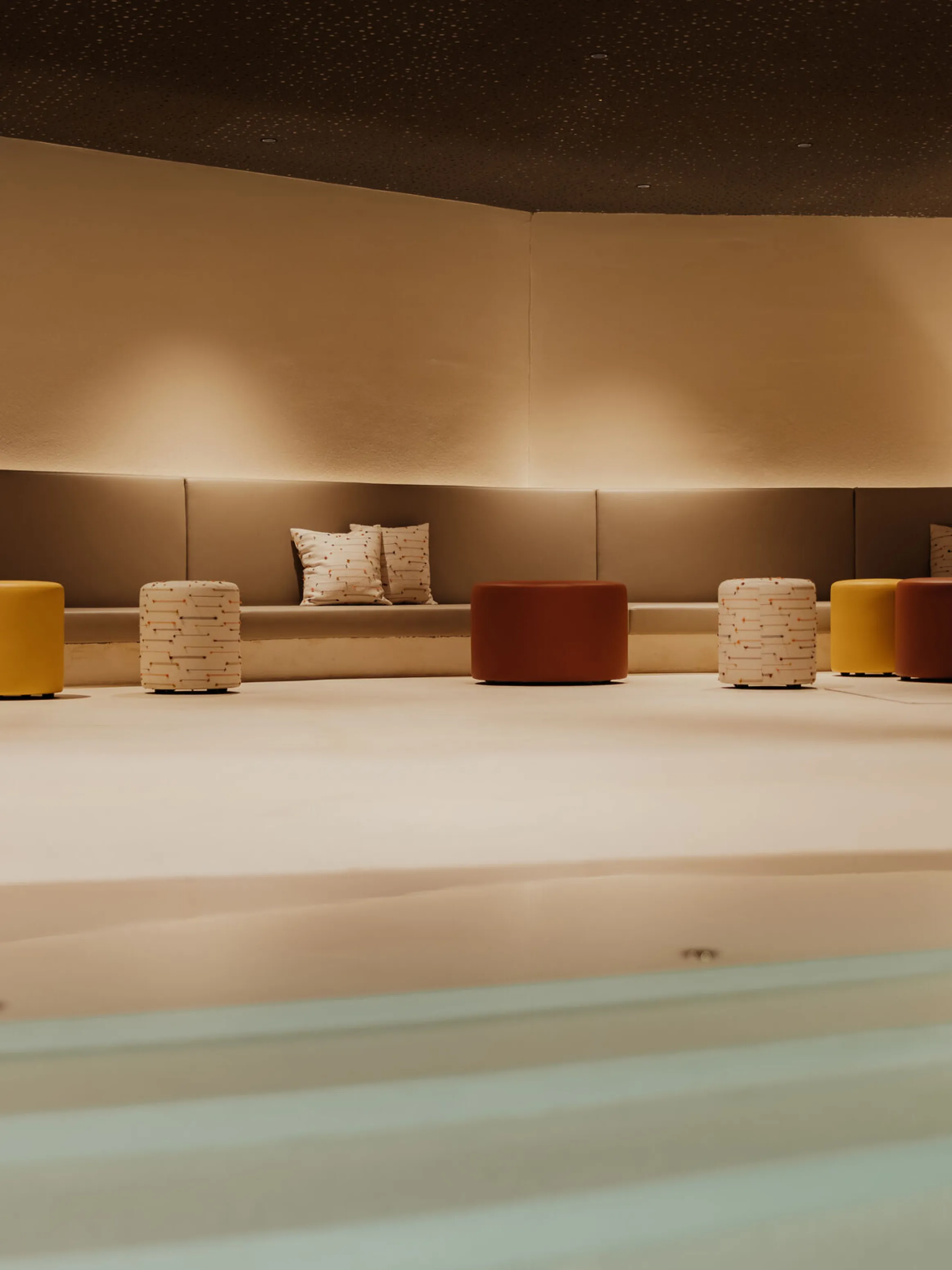 Modern lounge area with a cozy atmosphere. Features curved gray sofa, colorful ottomans in red, yellow, and white, and soft lighting. Calm and inviting.