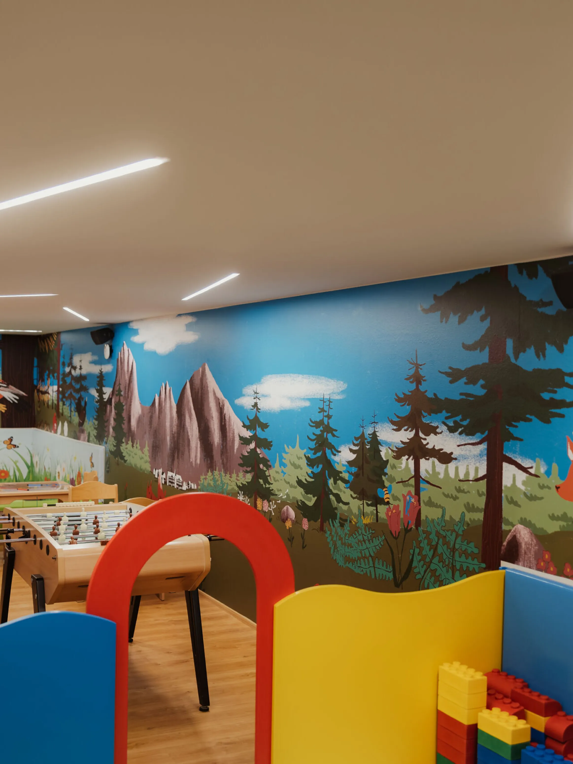 Colorful playroom with a vibrant forest-themed mural featuring trees and animals. Play tables, a foosball table, and building blocks create a playful atmosphere.