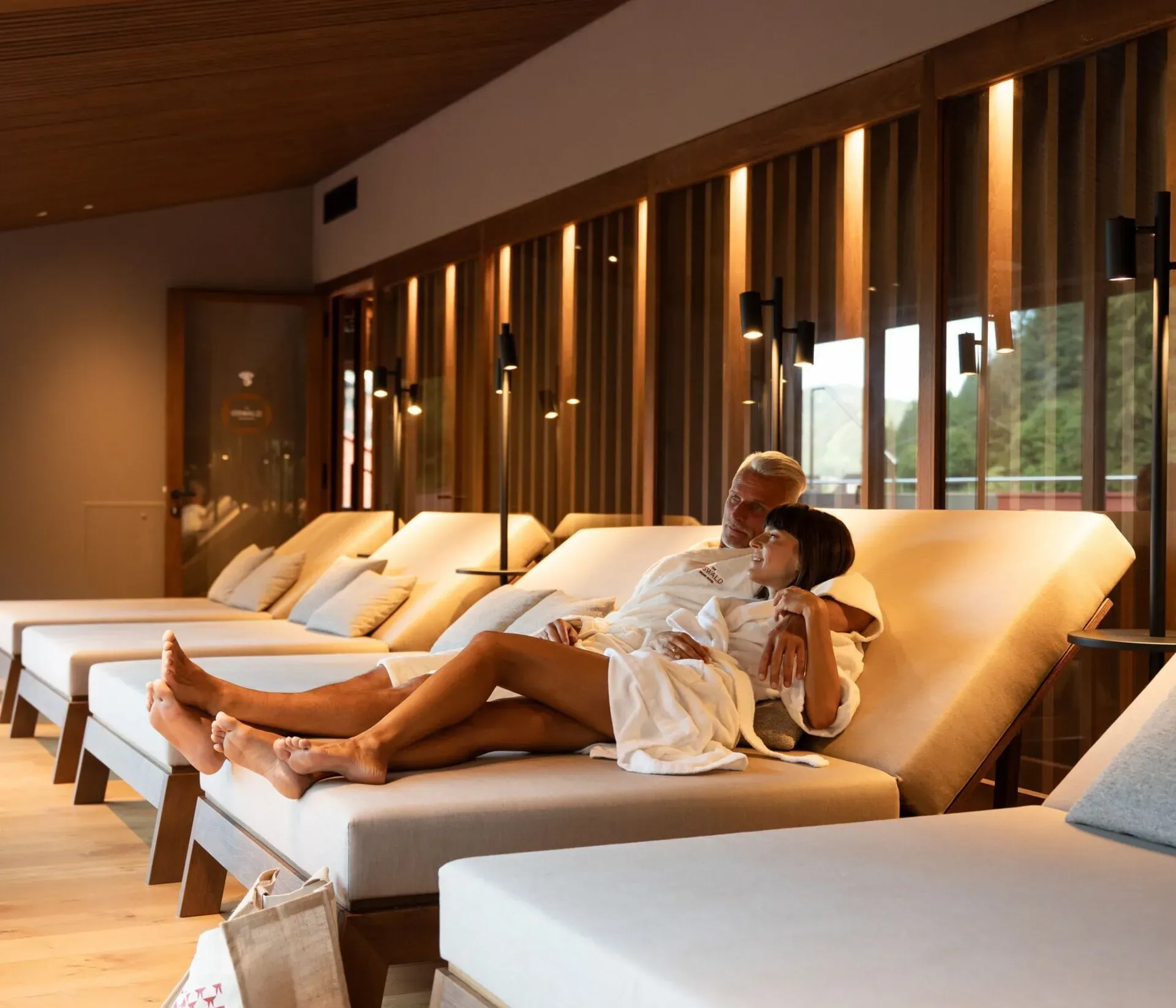 Elegant relaxation area at The Oswald Grand Hotel in Selva di Val Gardena, featuring comfortable ergonomically shaped loungers and warm indirect lighting. A couple wearing bathrobes relaxes together on one of the loungers. Natural wood, muted tones and soft lighting create a calm and welcoming atmosphere in the wellness area.