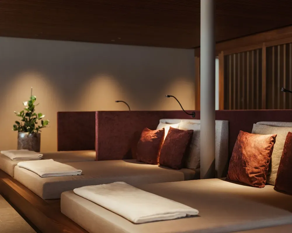 Comfortable relaxation area at The Oswald Grand Hotel with spacious upholstered loungers, folded towels and decorative cushions in natural and warm red tones. Small reading lamps and indirect lighting create a cosy atmosphere. Wooden details and plants complement the alpine-inspired interior design.