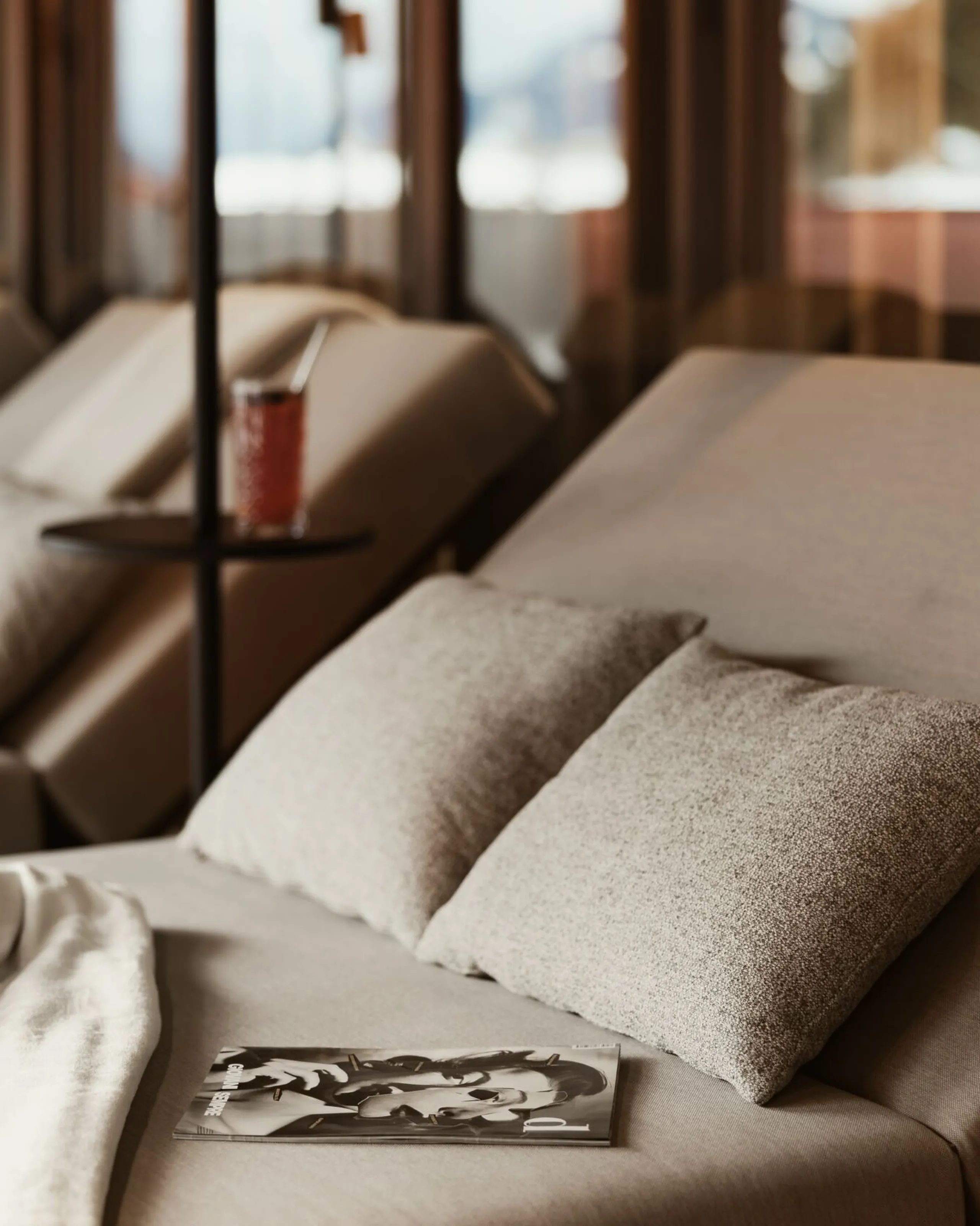 Close-up of a comfortable wellness lounger at The Oswald Grand Hotel. Two soft cushions and a neatly folded white towel are placed on the lounger. A magazine rests on a small side table. Additional loungers and warm wooden interior details can be seen softly in the background.