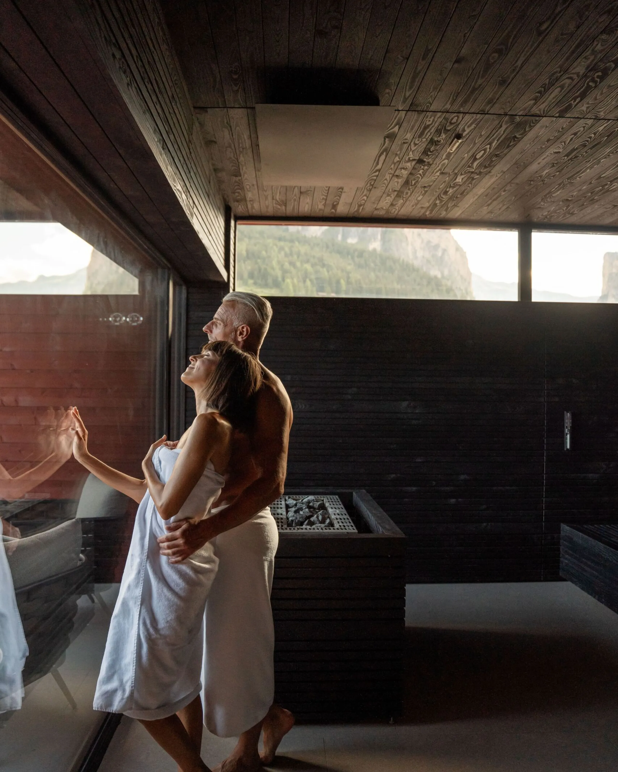 Couple sharing a peaceful moment in an elegant and intimate spa setting. The woman is wrapped in a white towel and leans against the man beside a large panoramic window. The interior features natural wood on the walls and ceiling, warm tones and natural materials. Part of the surrounding alpine landscape can be seen through the windows, creating a calm and private wellness atmosphere.
