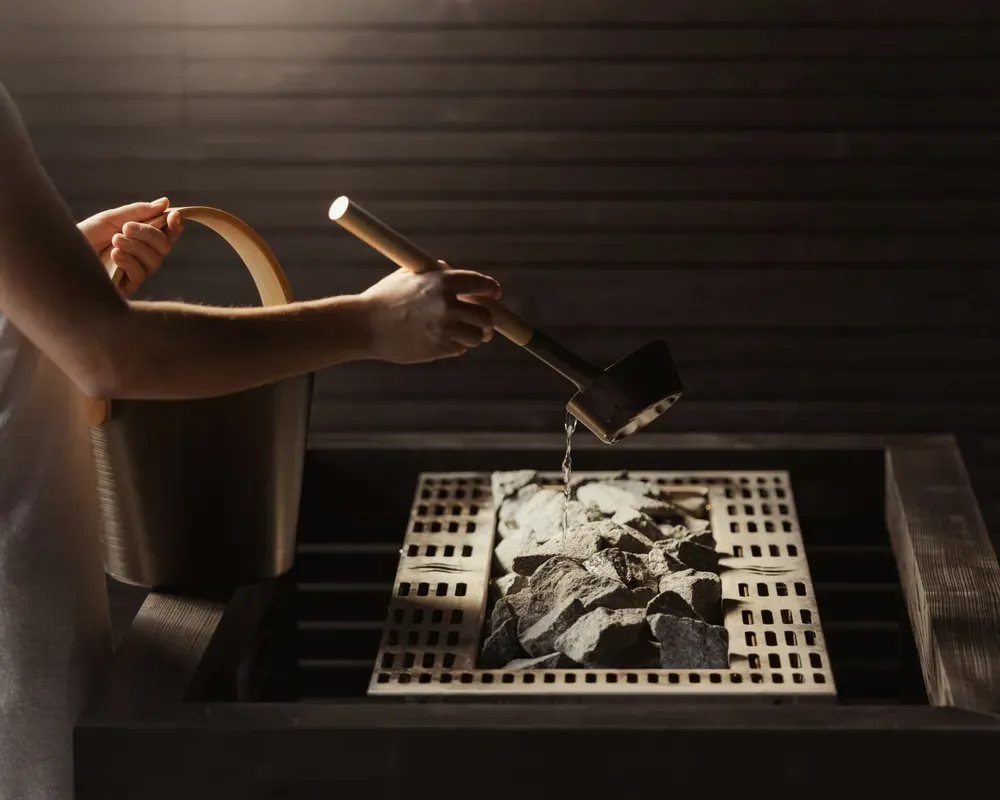 Close-up of a sauna infusion ritual in the wellness area of The Oswald Grand Hotel. A hand pours water over the hot sauna stones using a black ladle. A wooden bucket is positioned beside the sauna heater. Dark wood walls and warm lighting enhance the peaceful atmosphere of the sauna area.