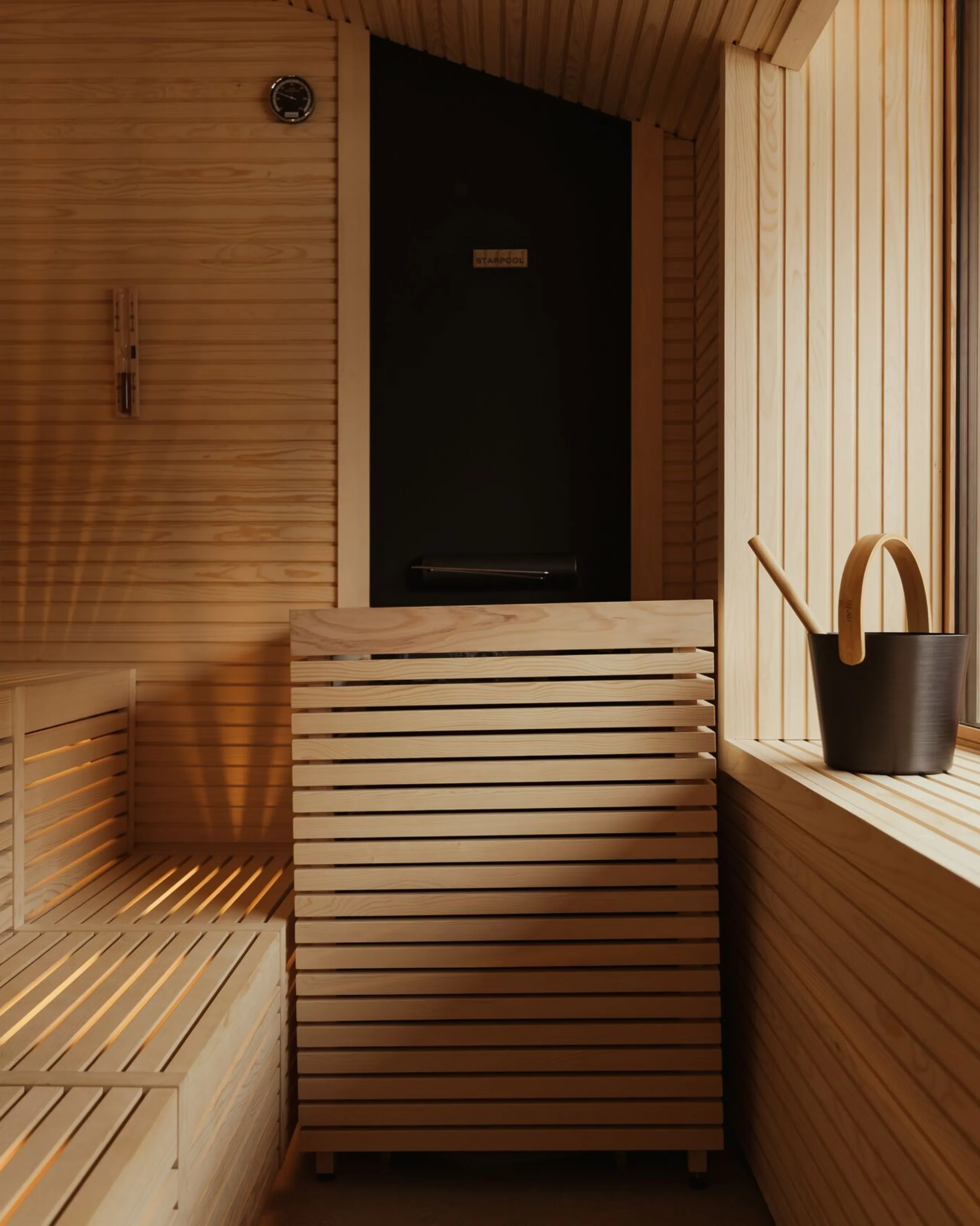 High-quality Bio sauna at The Oswald Grand Hotel, fully finished in light natural wood. The sauna features wide wooden benches and reclining surfaces, a modern sauna door and a large window allowing natural daylight into the space. A dark wooden bucket with a ladle for sauna infusions is positioned on the right.