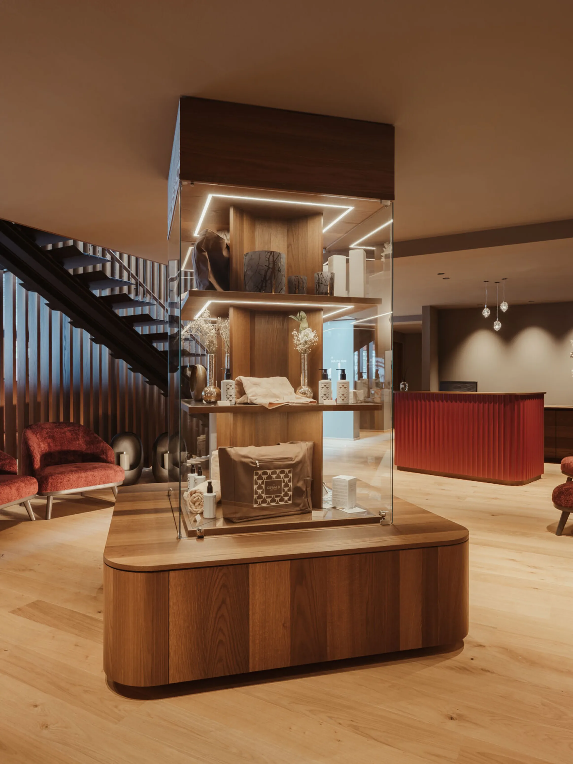 Elegant lobby with warm lighting, featuring a central wooden display case, plush red chairs, a modern staircase, and a sleek reception desk.