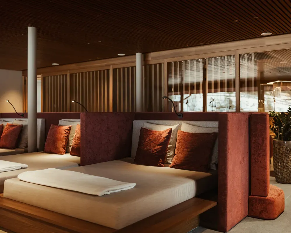Luxurious spa room with plush daybeds featuring rust-colored cushions, soft lighting, and wooden partitions. A potted plant adds greenery for relaxation.