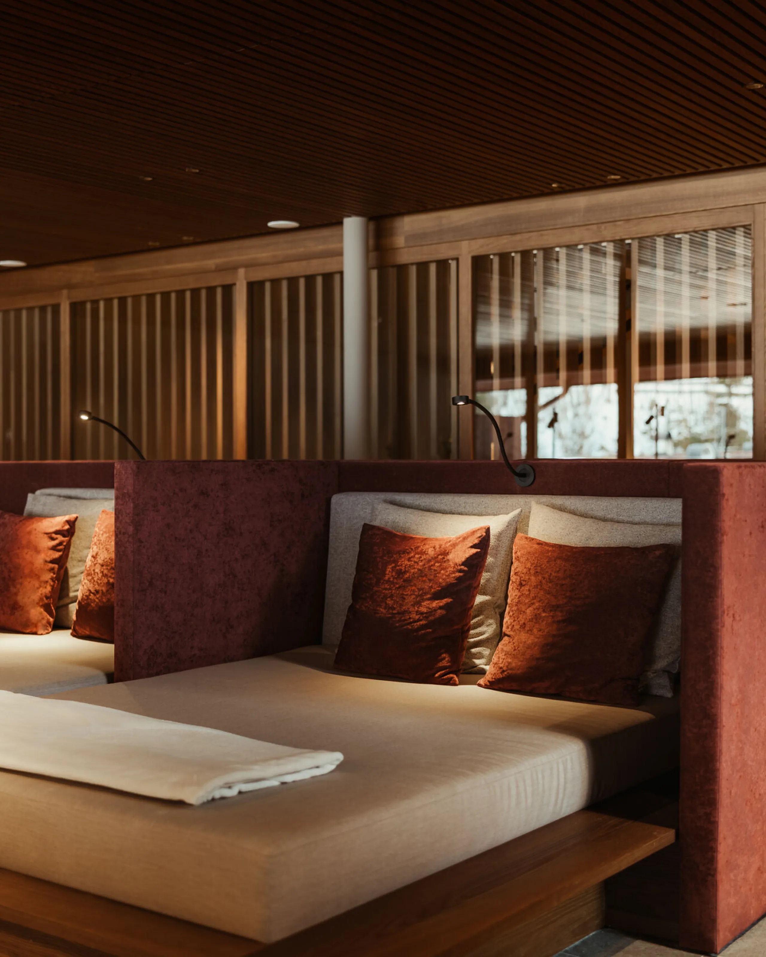 Stanza spa di lusso con lettini imbottiti dotati di cuscini color ruggine, illuminazione soffusa e divisori in legno. Una pianta in vaso aggiunge un tocco di verde per favorire il relax.