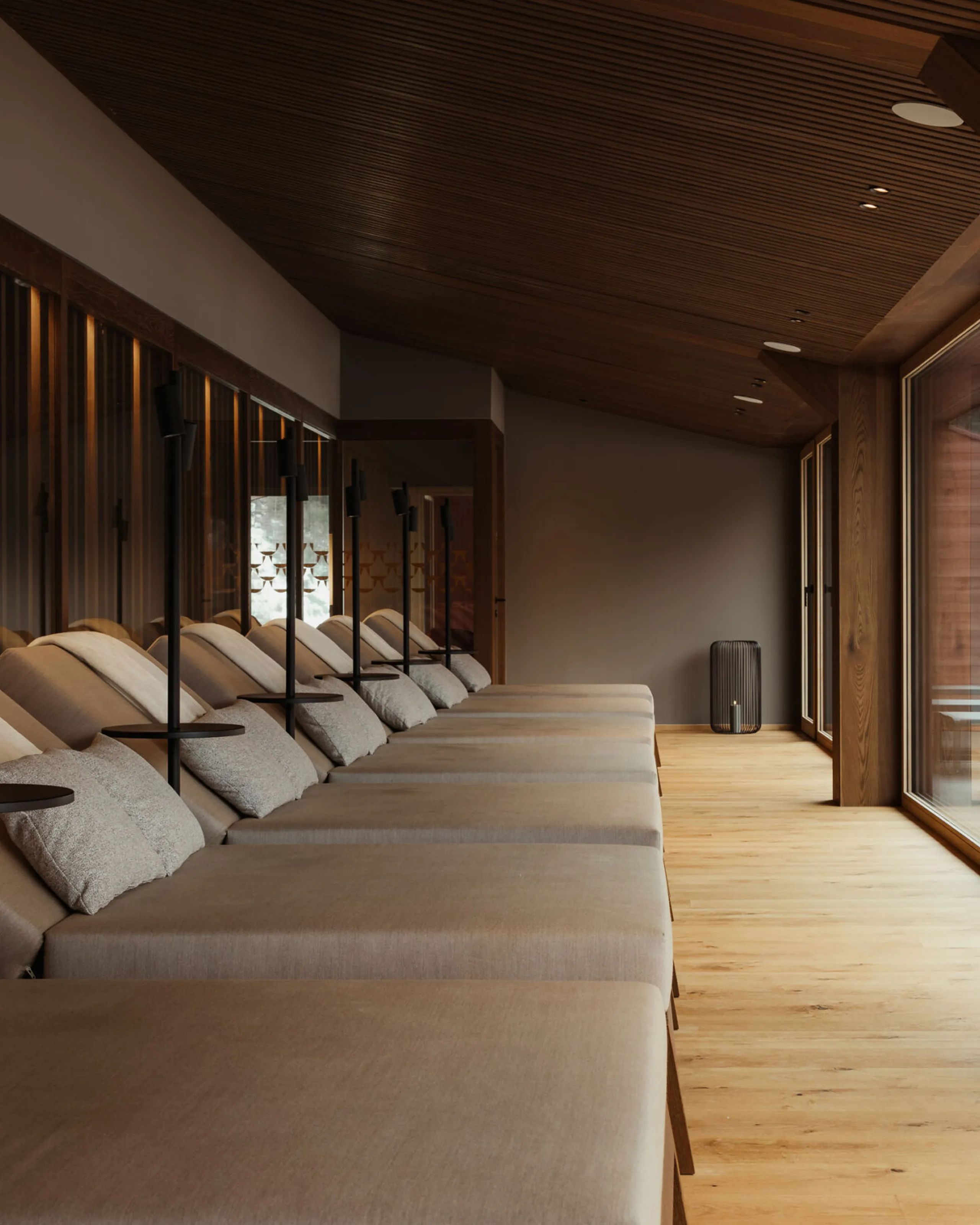 Moderner Spa-Relaxraum mit einer Reihe beiger Liegestühle, warmen Holztönen, großen Fenstern und ruhigen Ausblicken ins Freie, die eine entspannte Atmosphäre schaffen.