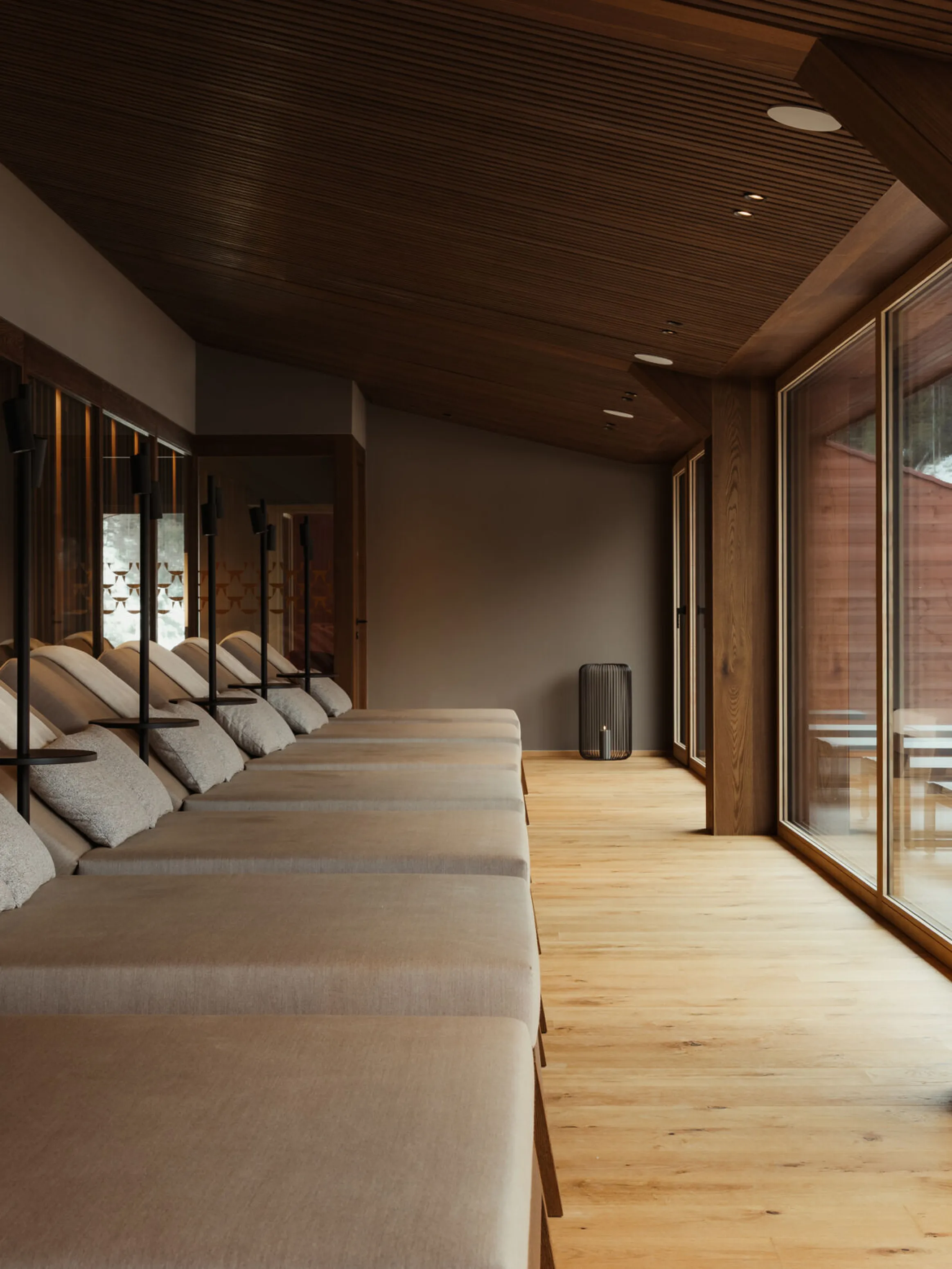 Modern spa relaxation room with a row of beige lounge chairs, warm wood tones, large windows, and serene outdoor views, creating a tranquil atmosphere.