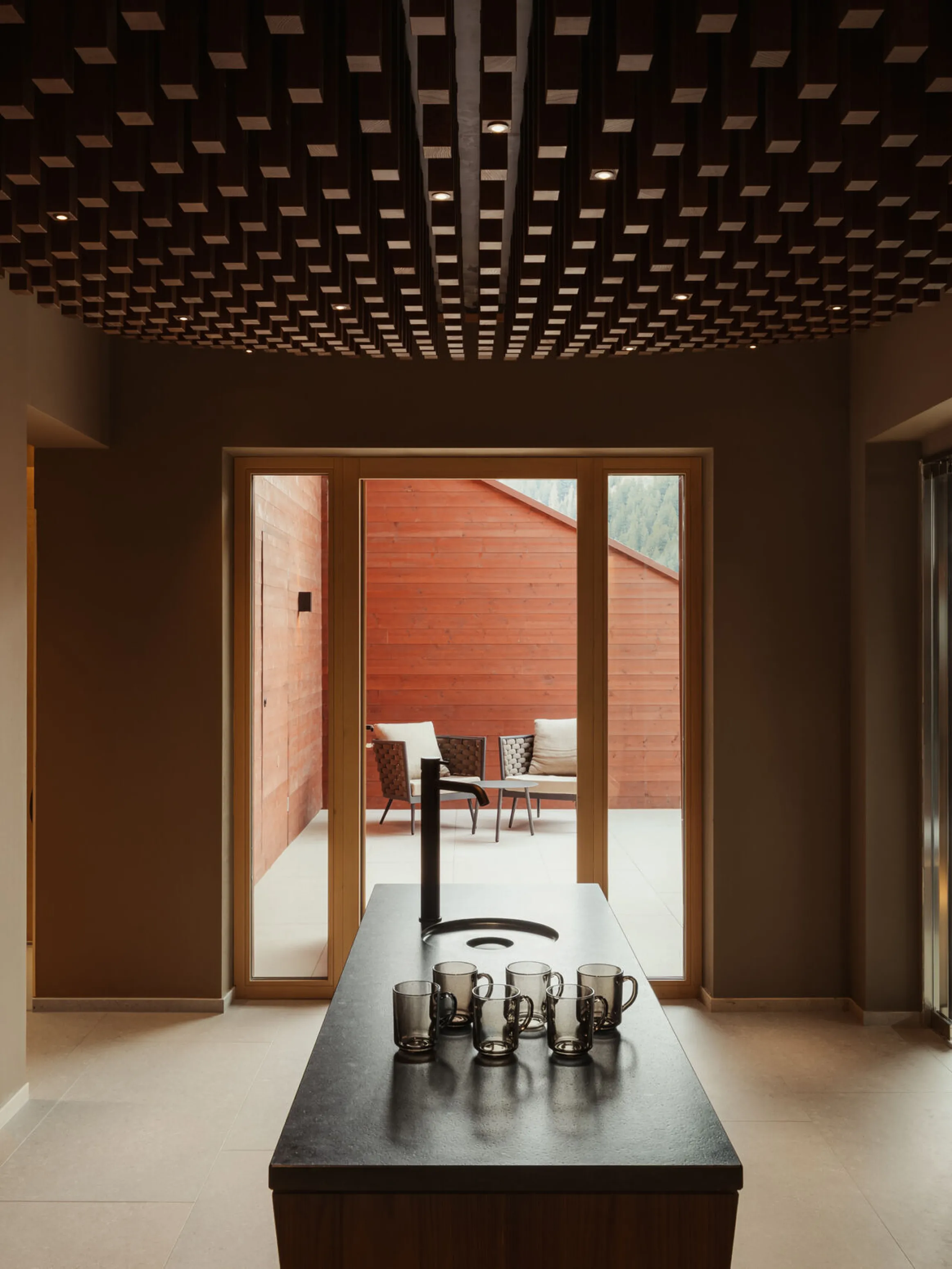 A modern interior with a dark wooden ceiling and a minimalist black table with several cups. Sunlit patio and seating are visible through large glass doors.