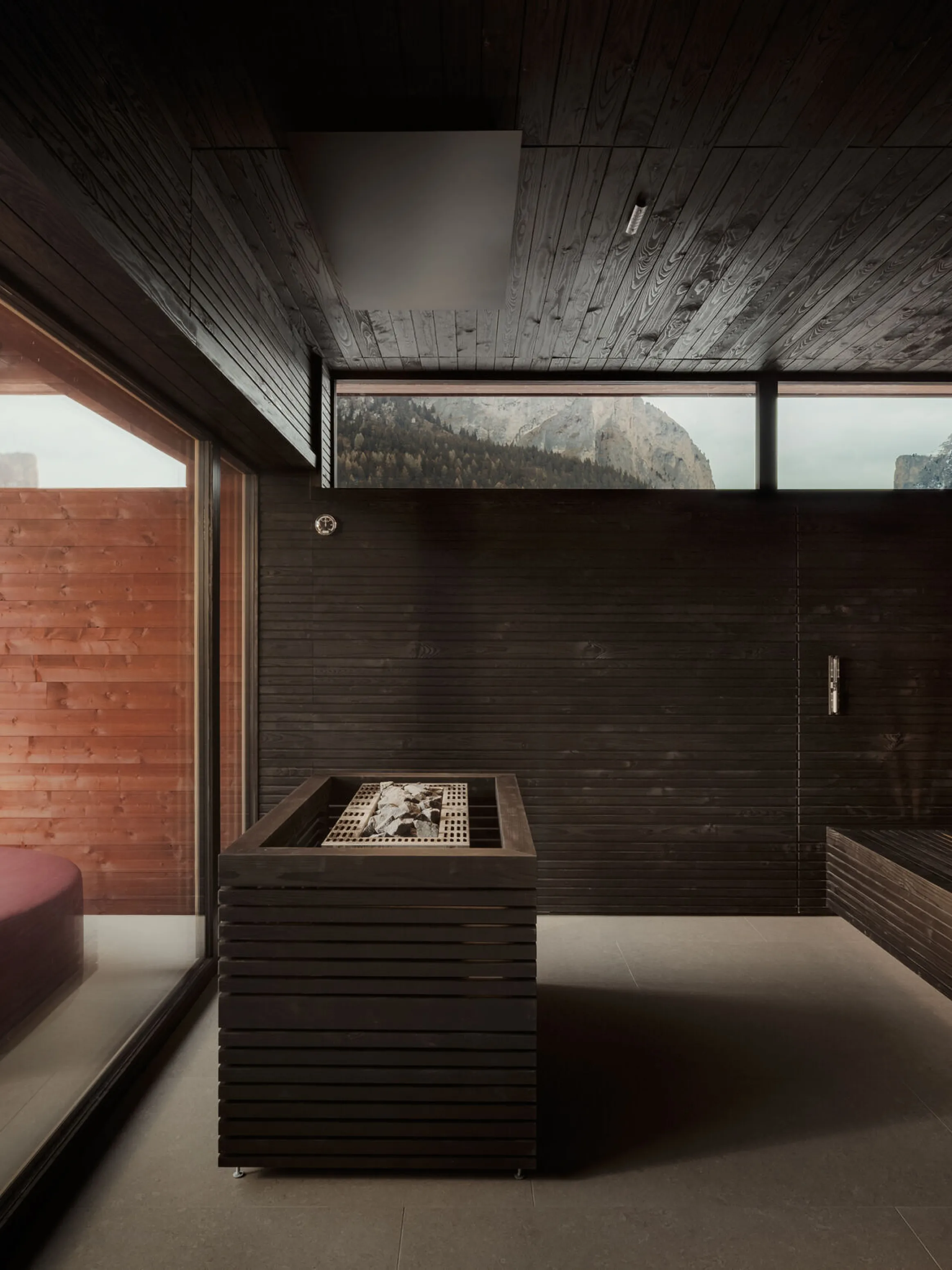 Modern sauna interior with dark wooden walls and tiered seating. Large windows reveal snowy mountain view. Warm tones create a serene, cozy atmosphere.