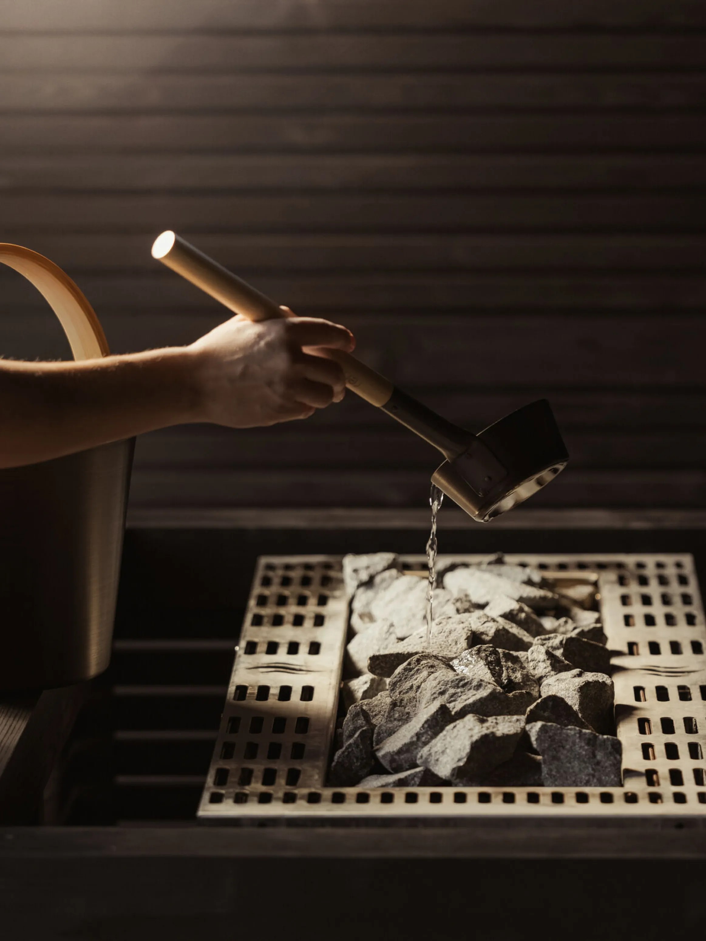 A person wearing a towel pours water from a ladle over hot sauna stones, creating steam. The setting is a dimly lit wooden sauna, evoking relaxation.