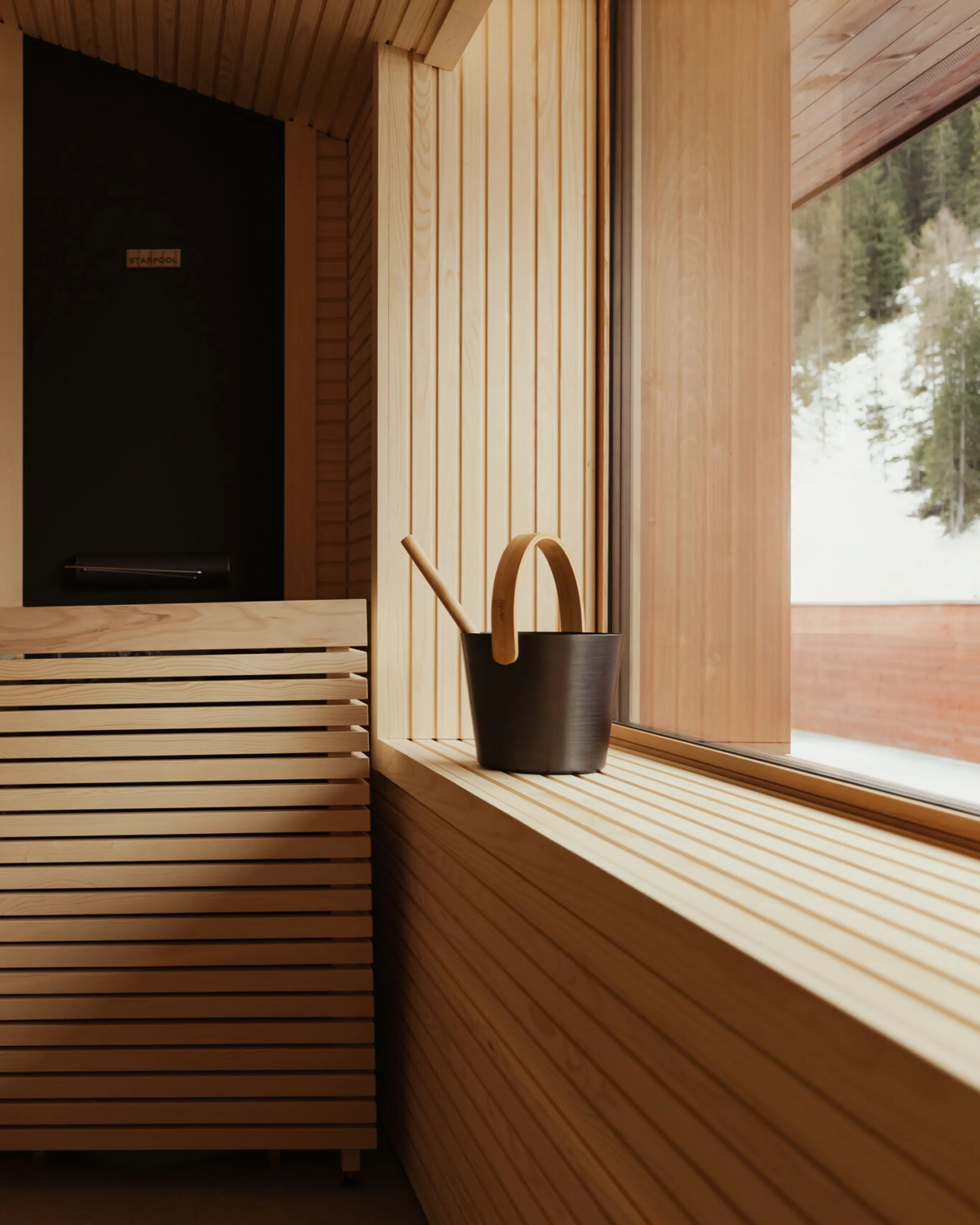 Modernes Holzsauna-Innenraum mit lamellenförmigen Wänden und Bank. Eine Gießkanne steht auf einer breiten Fensterbank, draußen sind schneebedeckte Bäume zu sehen. Ruhige Atmosphäre.