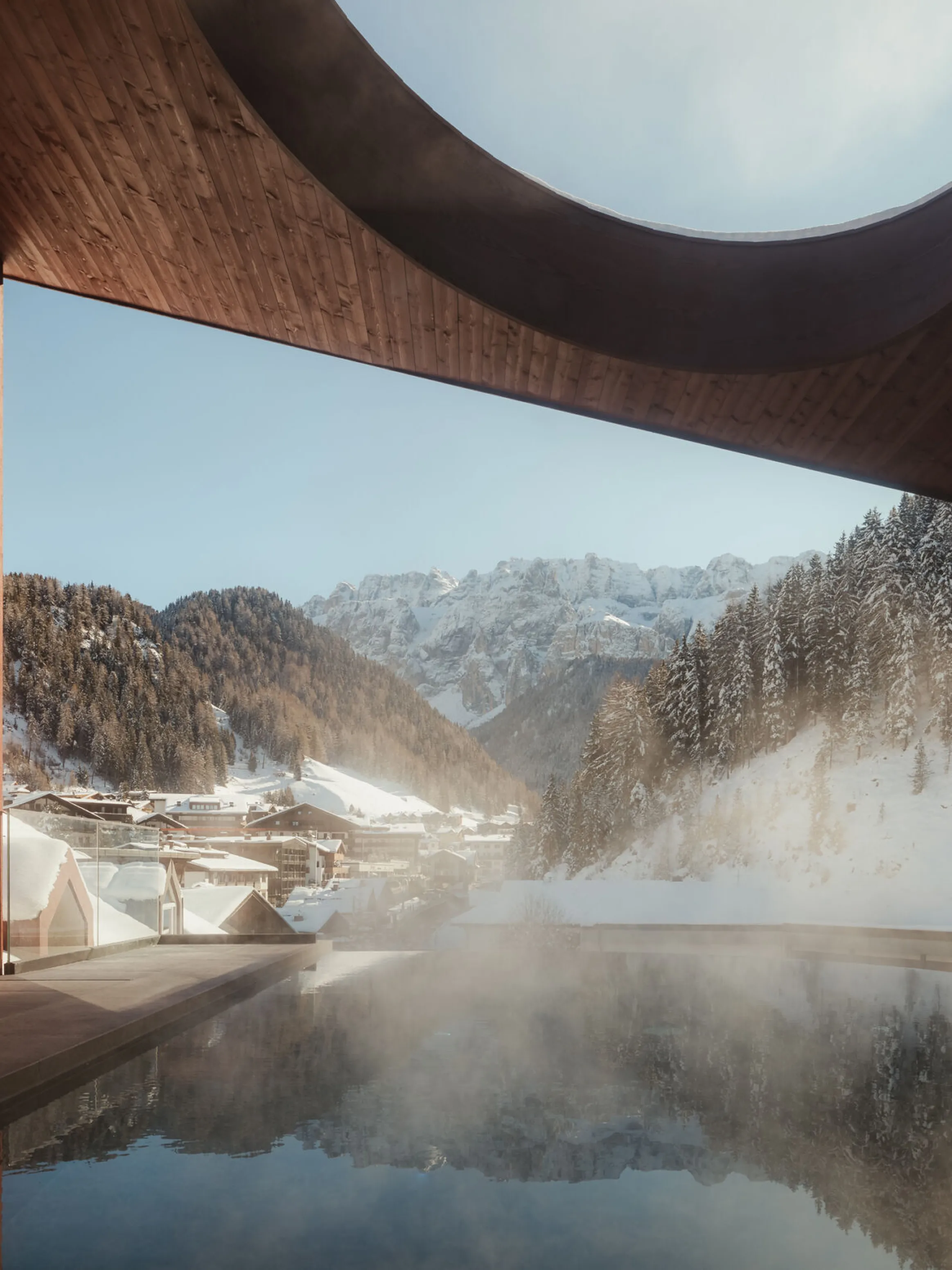 Modern architecture frames a snowy mountain village, seen from a steamy pool with a circular skylight, conveying tranquility and contrast.