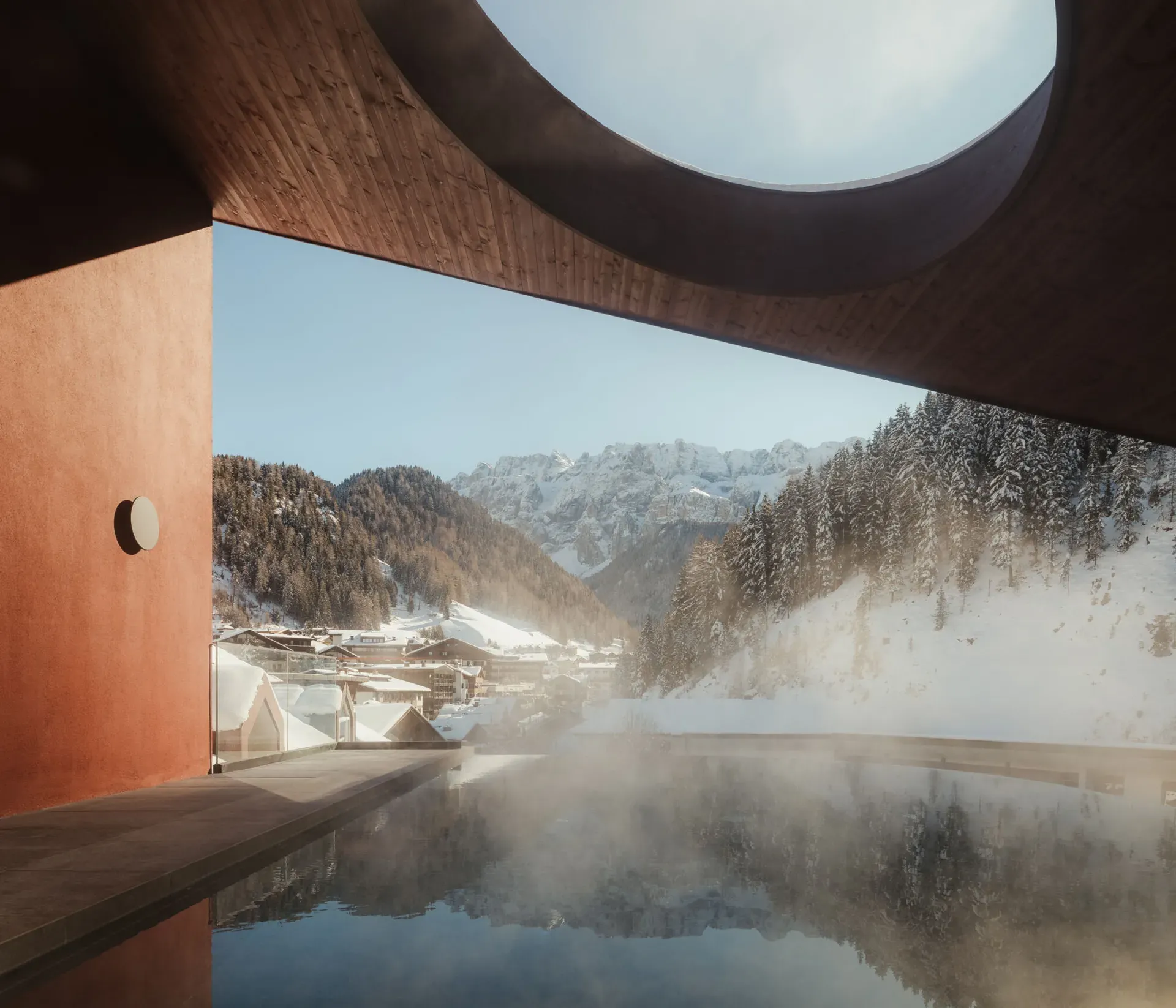 Modern architecture frames a snowy mountain village, seen from a steamy pool with a circular skylight, conveying tranquility and contrast.