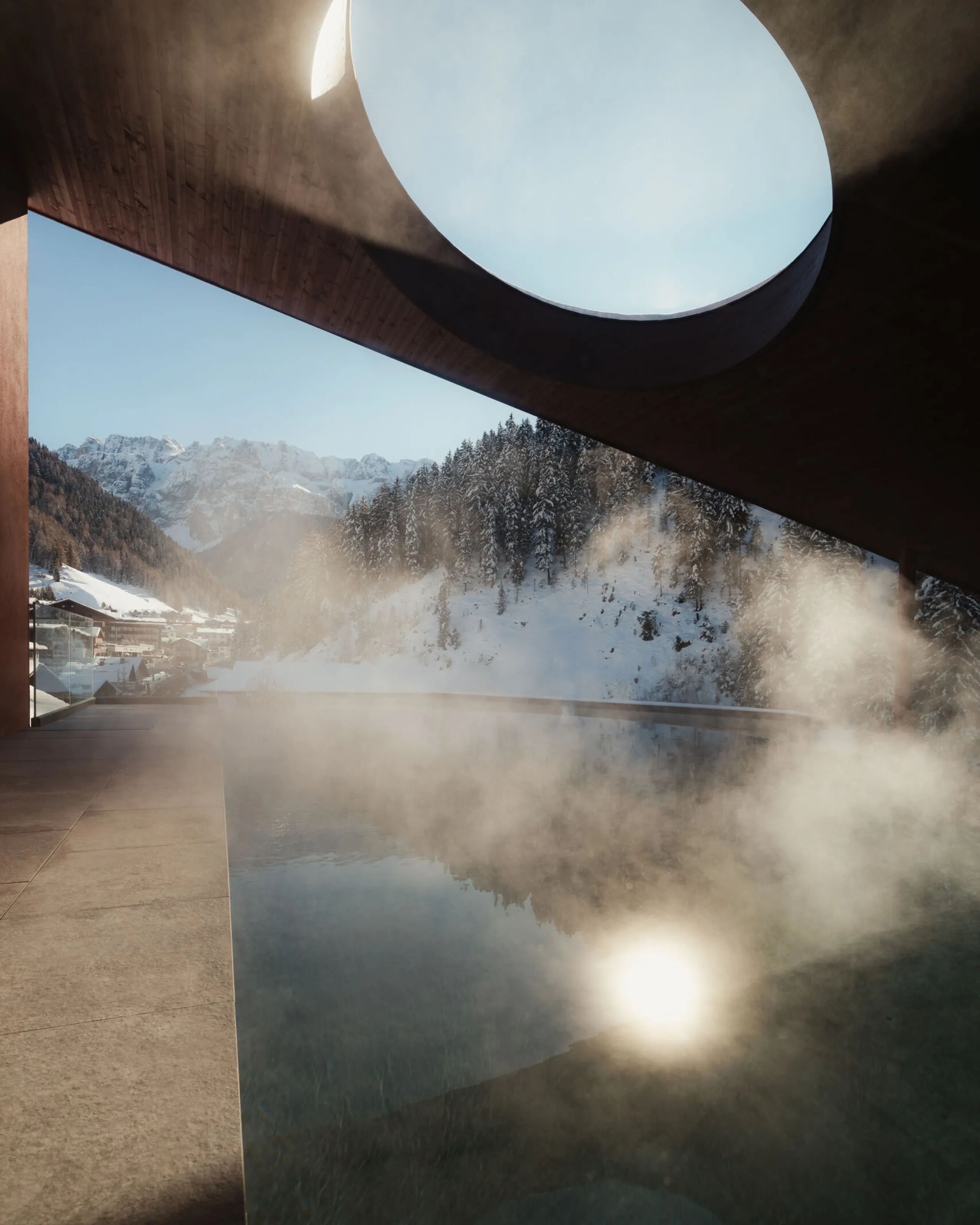 A circular opening in a modern roof casts light onto a steaming infinity pool, with snowy mountains and pine trees creating a serene winter backdrop.