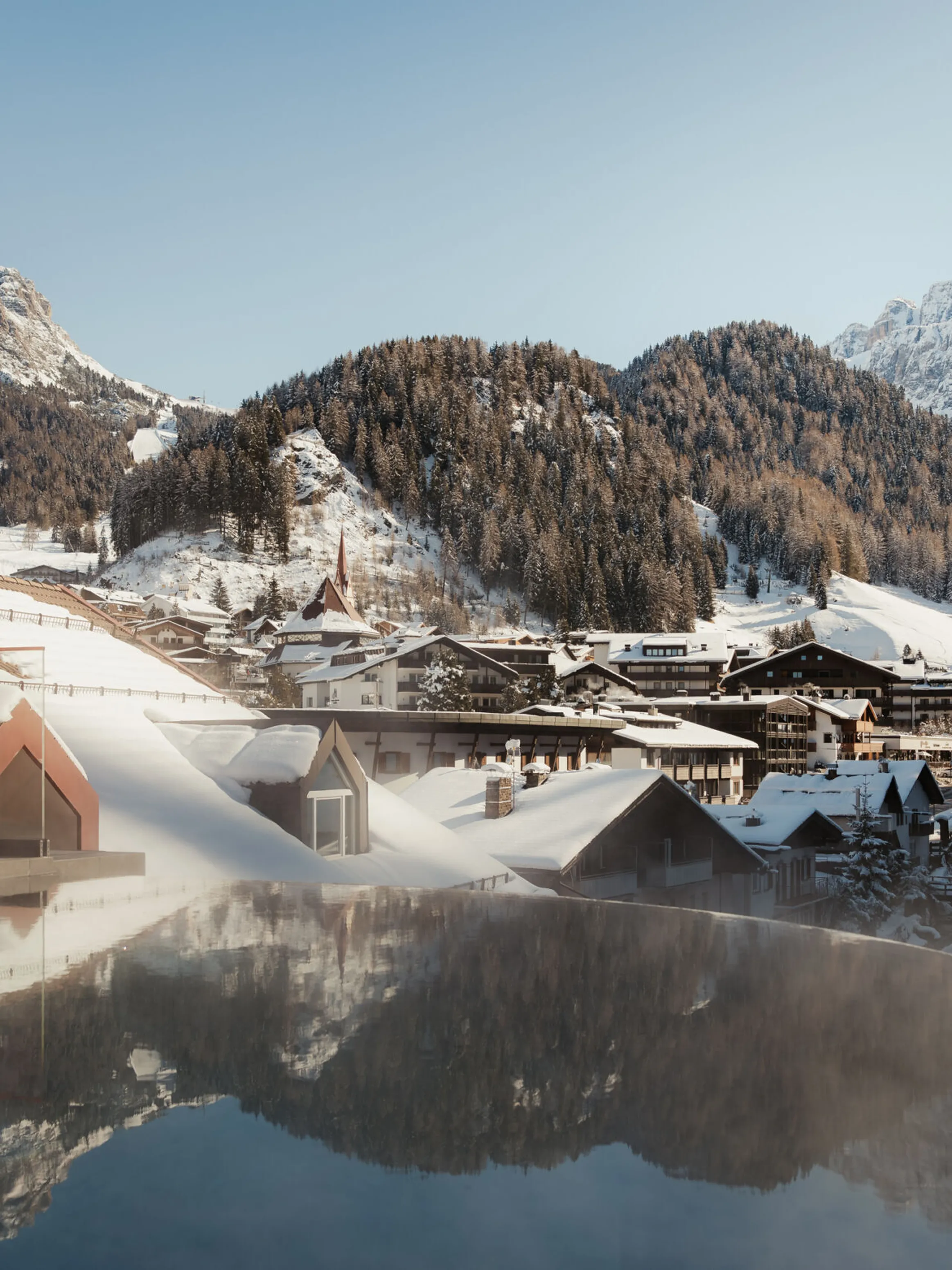 Snow-covered mountain village with wooden houses under a blue sky. Steaming rooftop pool in the foreground adds a serene contrast to the icy landscape.