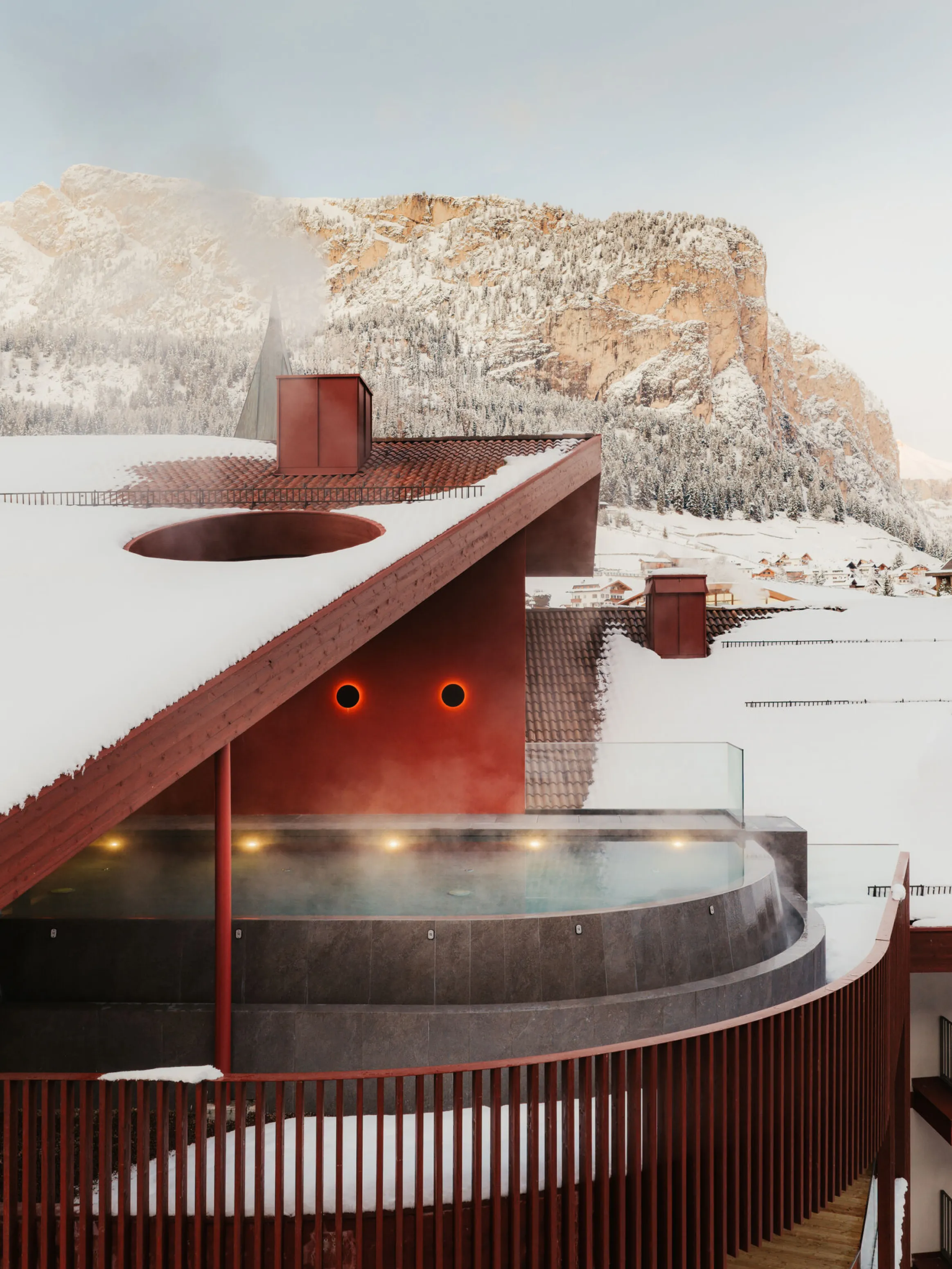 A modern building with red accents and snow-covered roof, featuring a steaming outdoor pool. In the background, snow-capped mountains under a clear sky.