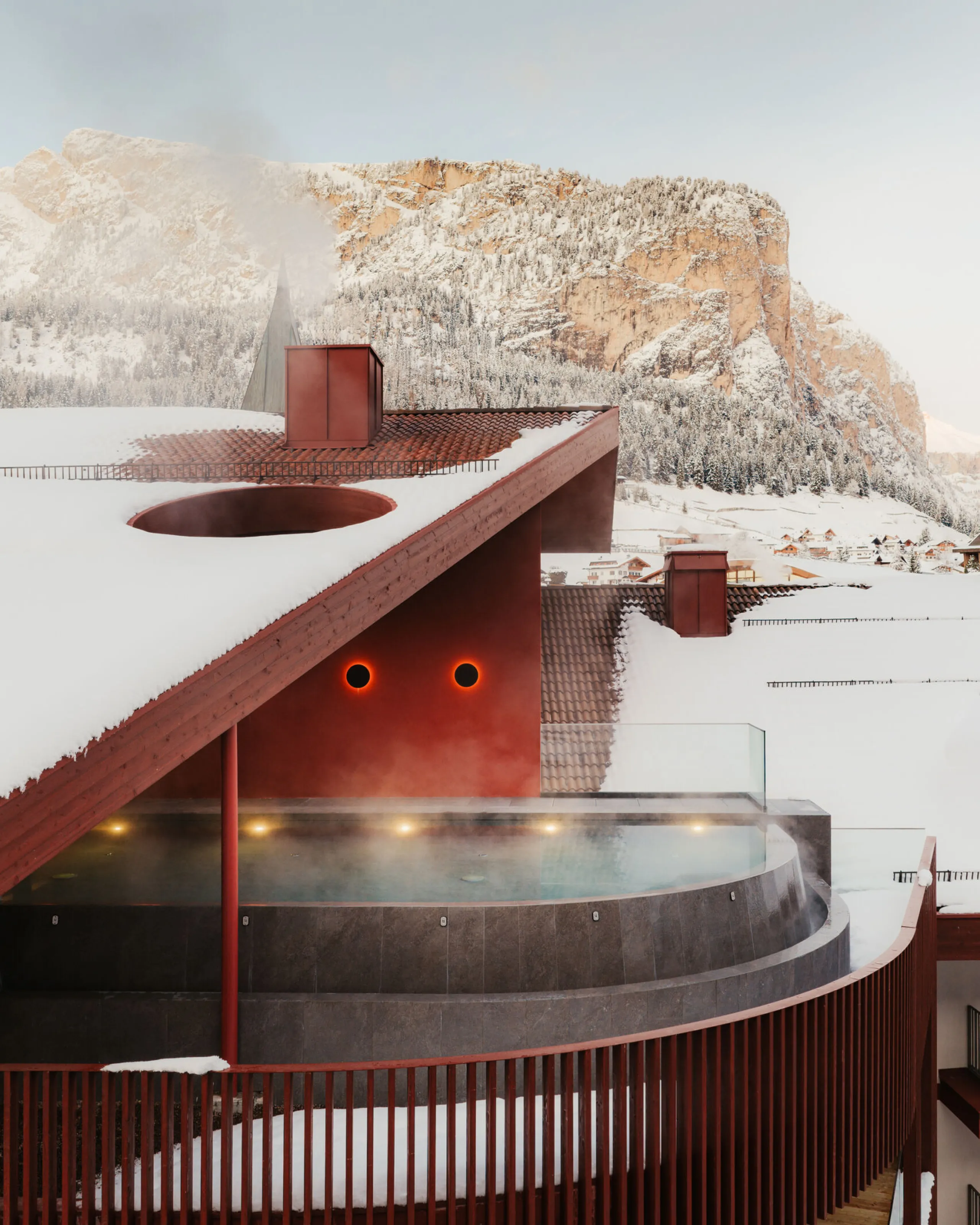 A modern building with red accents and snow-covered roof, featuring a steaming outdoor pool. In the background, snow-capped mountains under a clear sky.