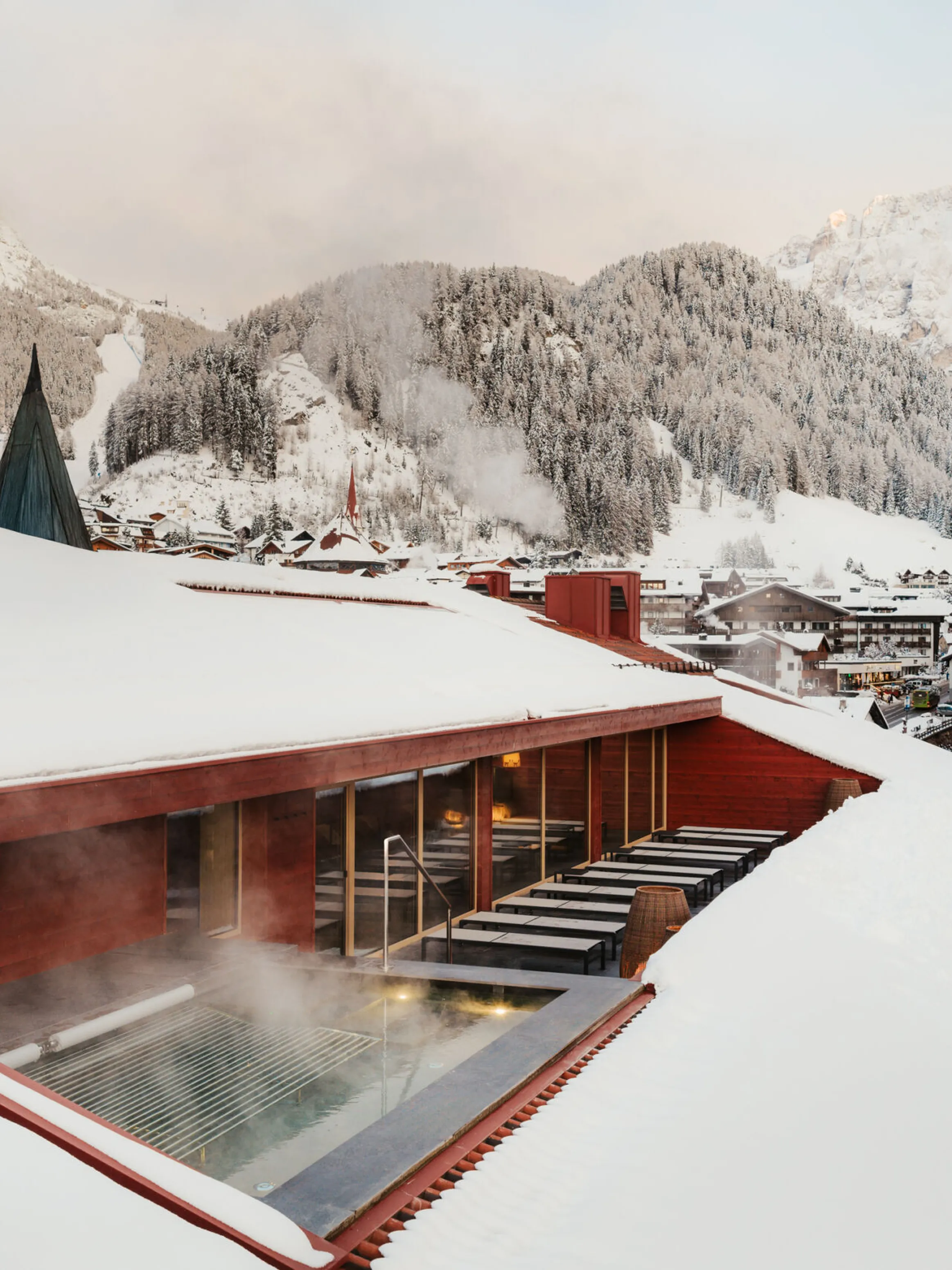 A snow-covered rooftop surrounds a steaming outdoor pool, with a picturesque view of snowy mountains and a cozy village nestled below, evoking calmness.