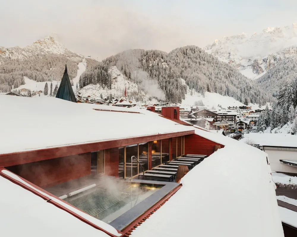 A snow-covered rooftop surrounds a steaming outdoor pool, with a picturesque view of snowy mountains and a cozy village nestled below, evoking calmness.