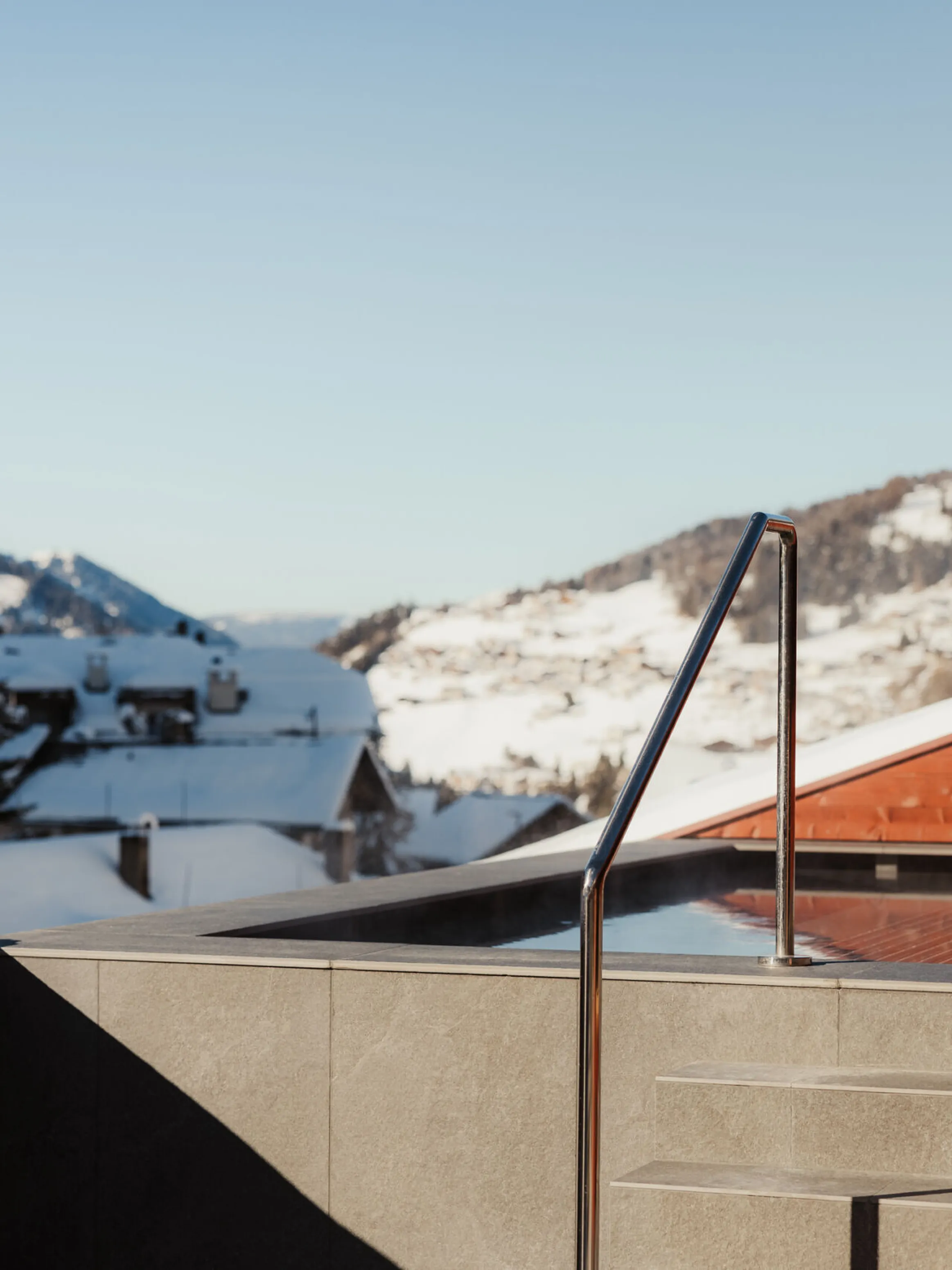 Rooftop hot tub with steam rising, set against a snowy alpine village and mountains under a clear blue sky, conveying tranquility and luxury.